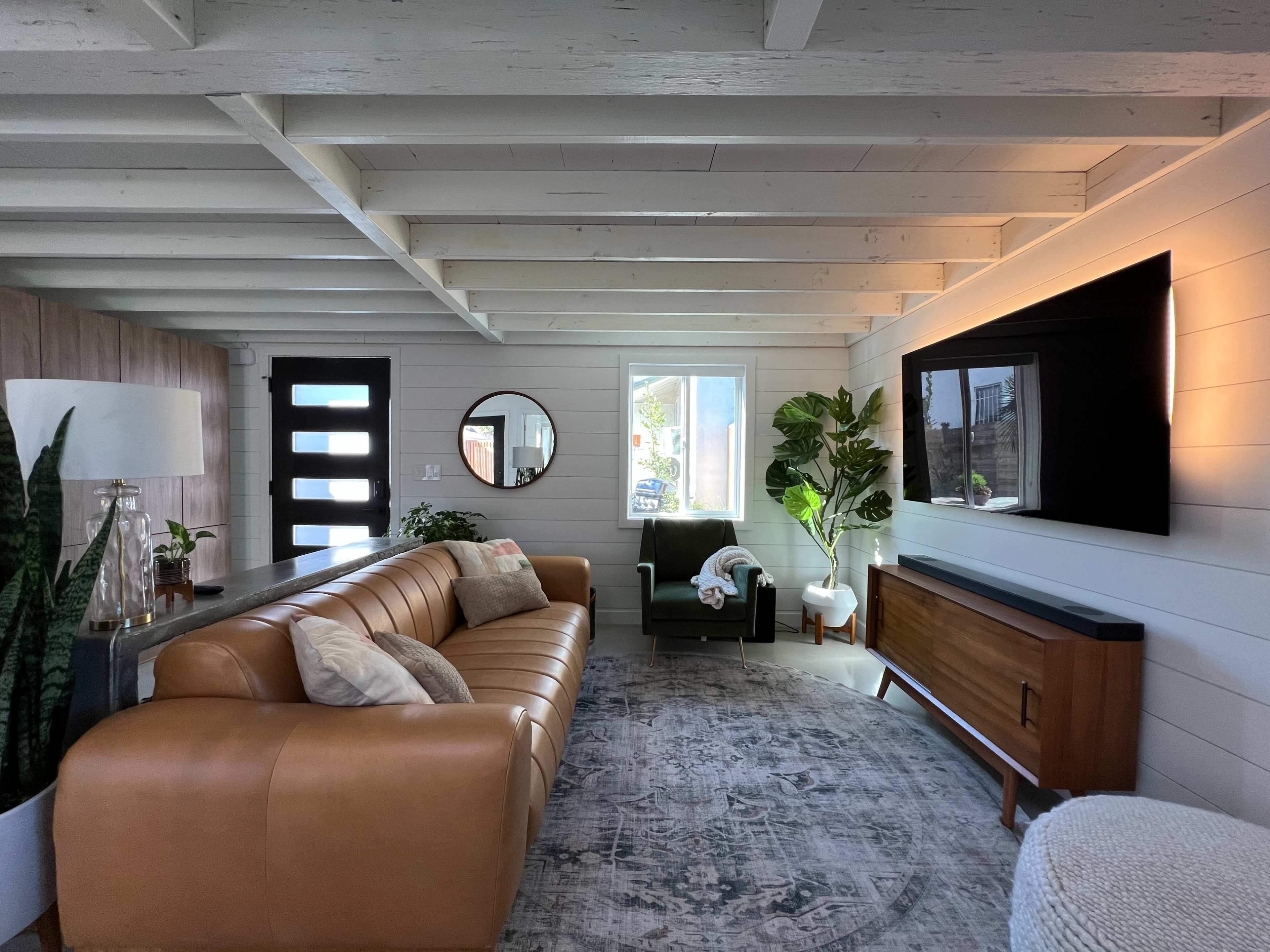 The living room features a tan leather sofa, a dark green armchair, a wooden media console beneath a wall-mounted TV, and a large window beside a round mirror.