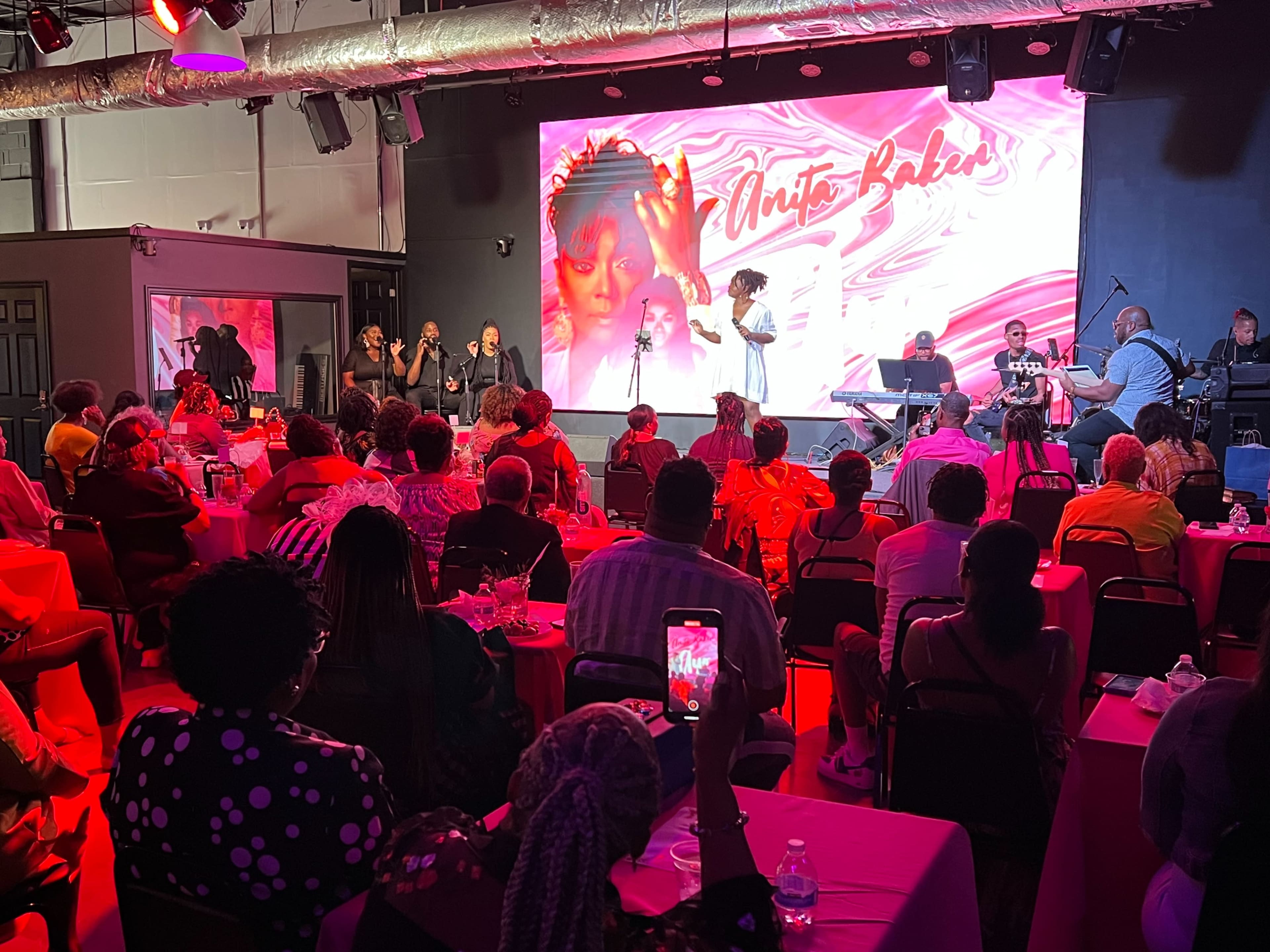 A performance event featuring a singer on stage with a band, while an audience sits at tables illuminated by red lighting.