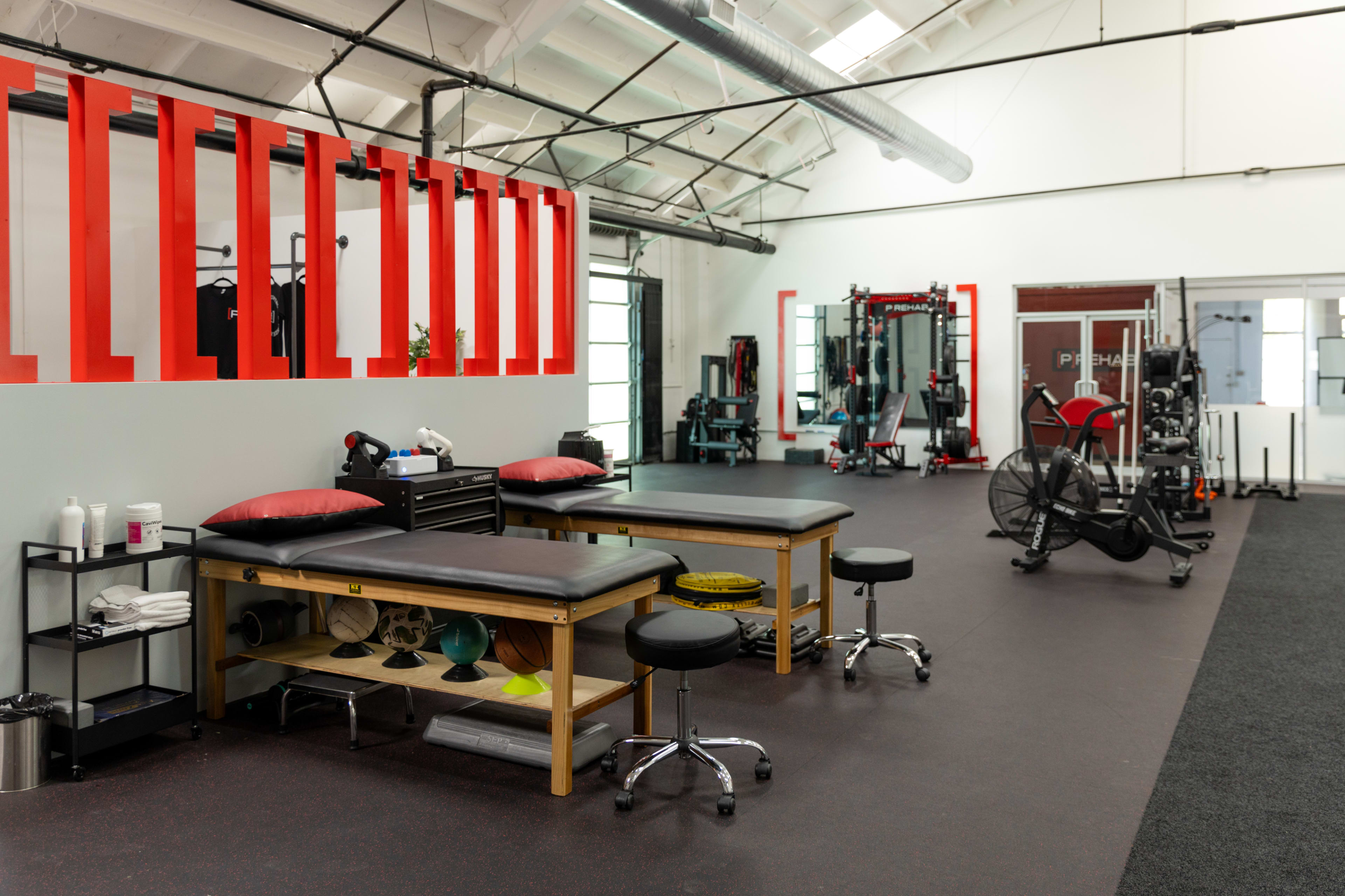 The image shows a fitness therapy room with two treatment tables, exercise equipment, and workout machines in a spacious, well-lit environment.