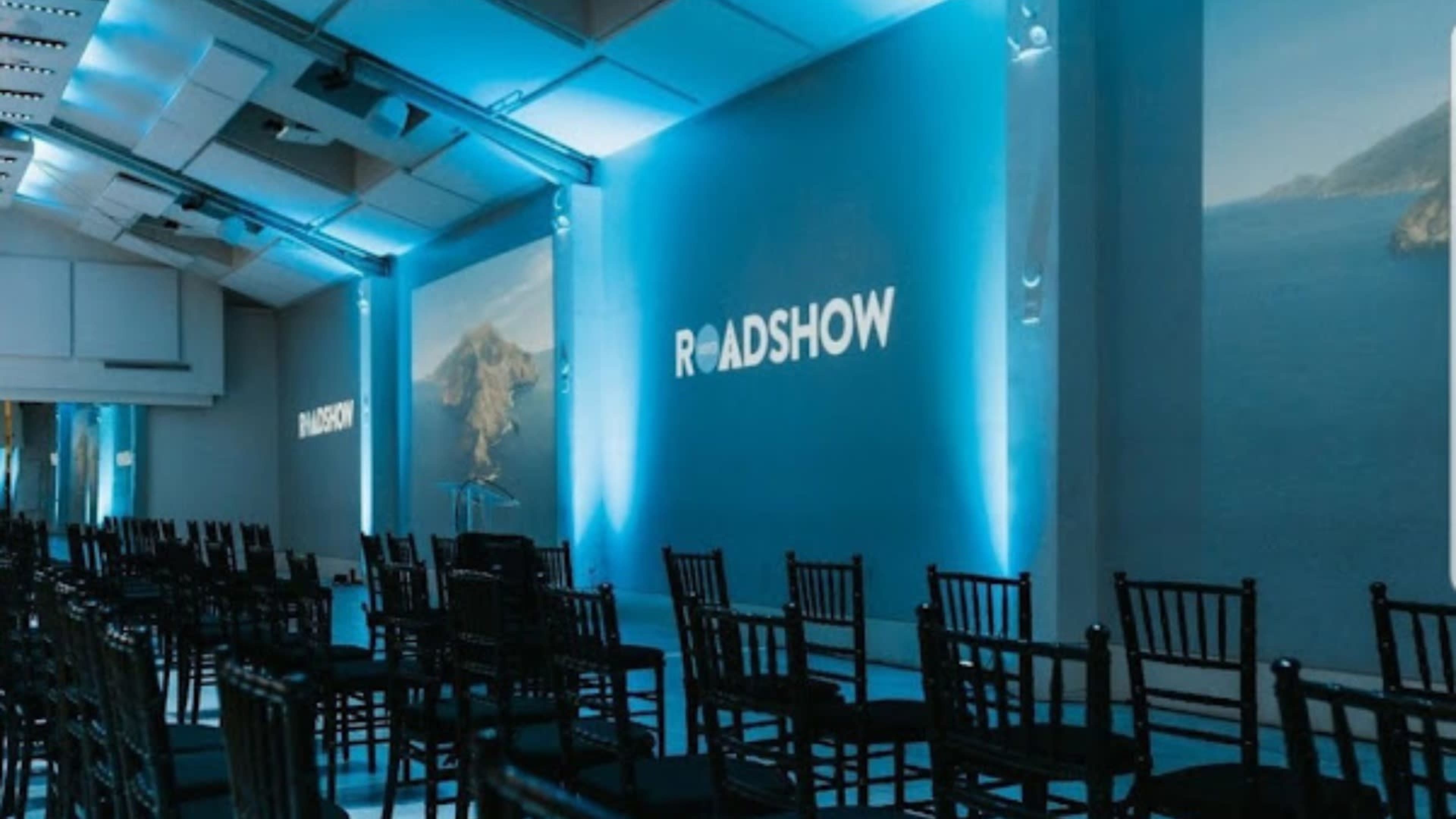 The image shows a room set up for an event, featuring rows of black chairs facing large screens displaying the word "ROADSHOW," with a blue light illuminating the space.