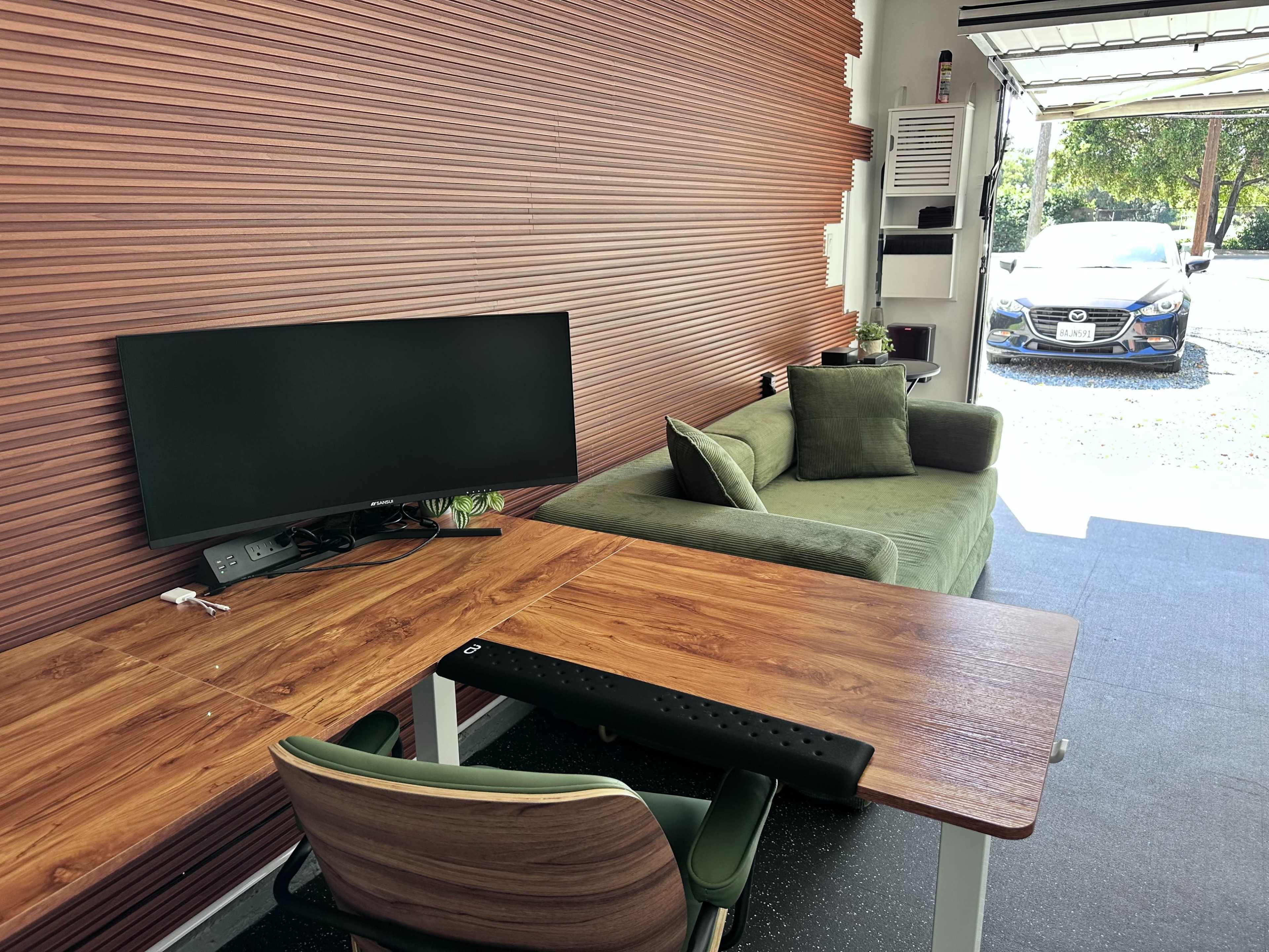 The image shows a modern workspace featuring a wooden desk with a green chair, a television mounted on the wall, and a green sofa, with a view of a car parked outside through a garage door.