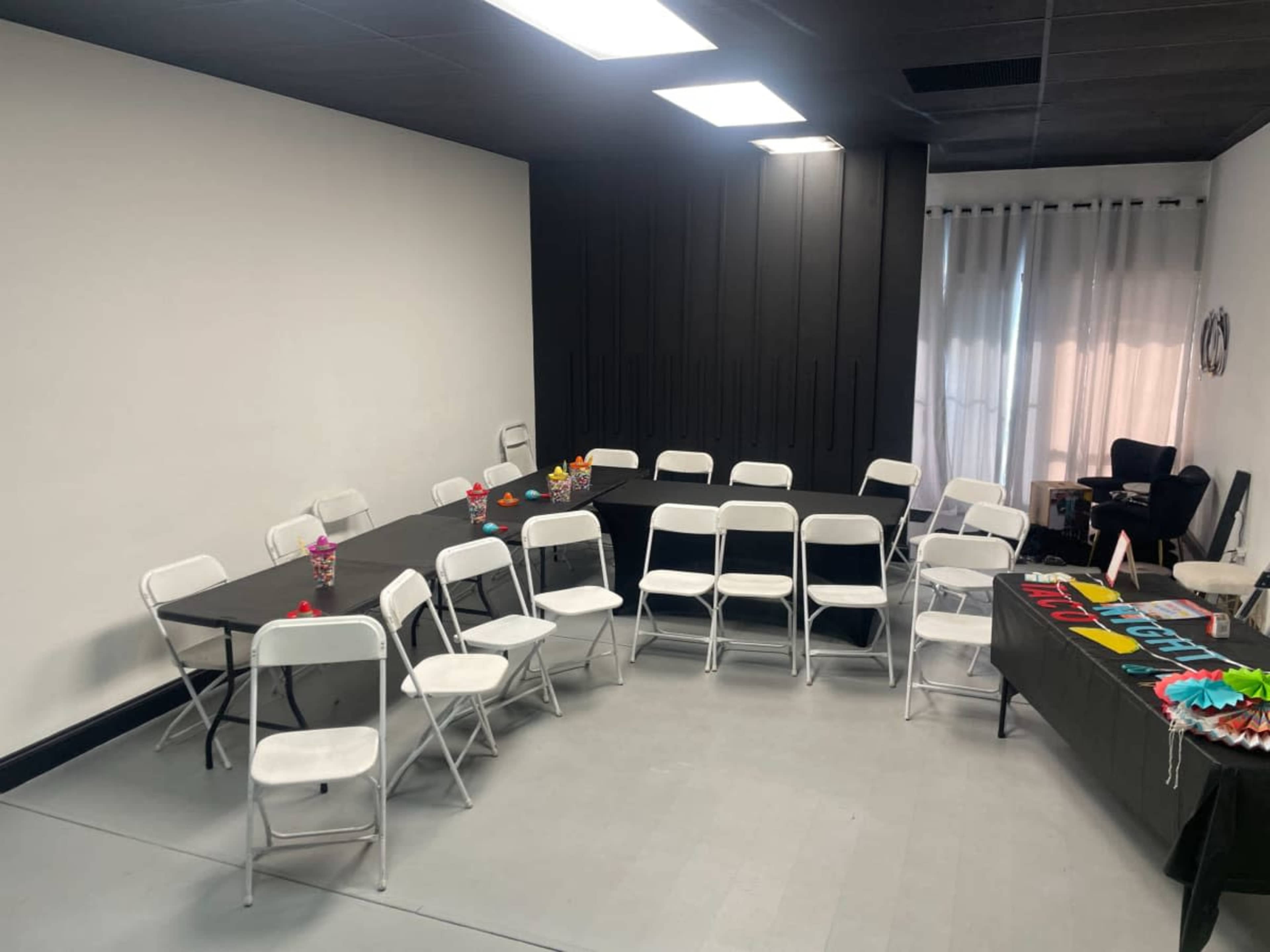 The image shows a meeting room with several white folding chairs arranged around tables, a window with curtains, and a black wall cabinet.