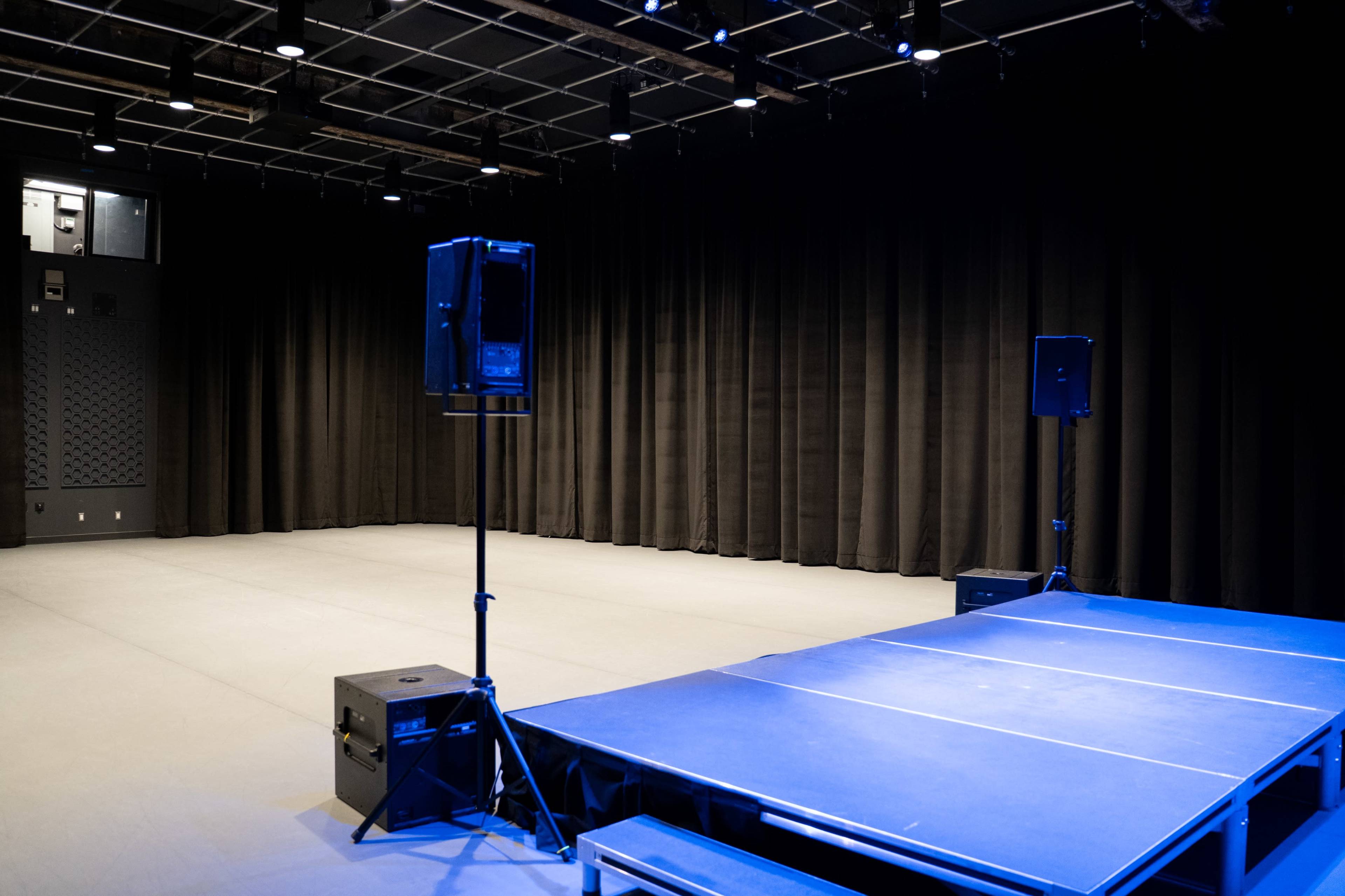 The image shows an empty performance stage with a blue lighting setup, flanked by two speaker stands, and black curtains in the background.