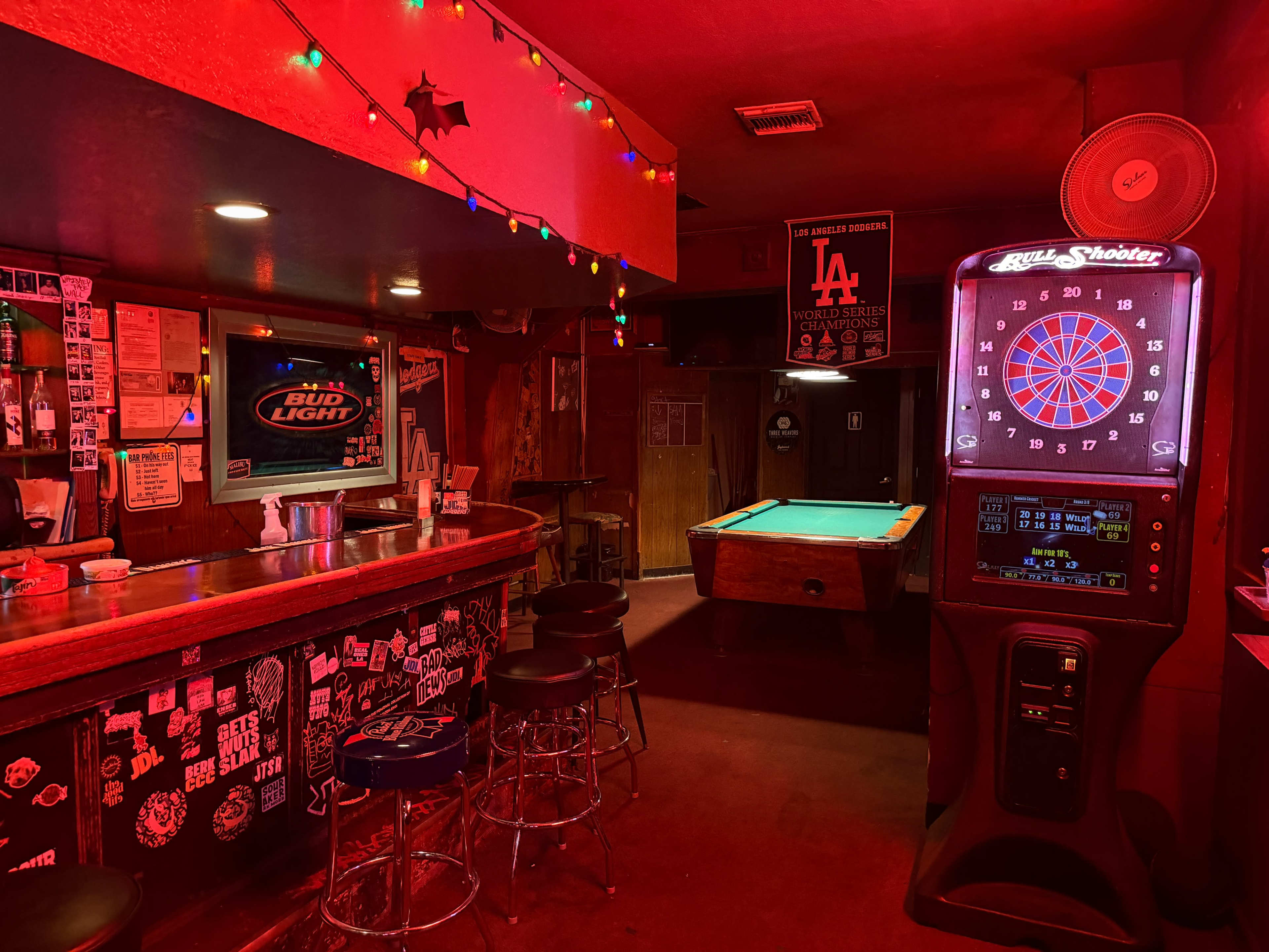 The image shows a dimly lit bar with a pool table, a dart machine, a television, and a wooden counter, all illuminated by red lighting and decorated with various posters and stickers.