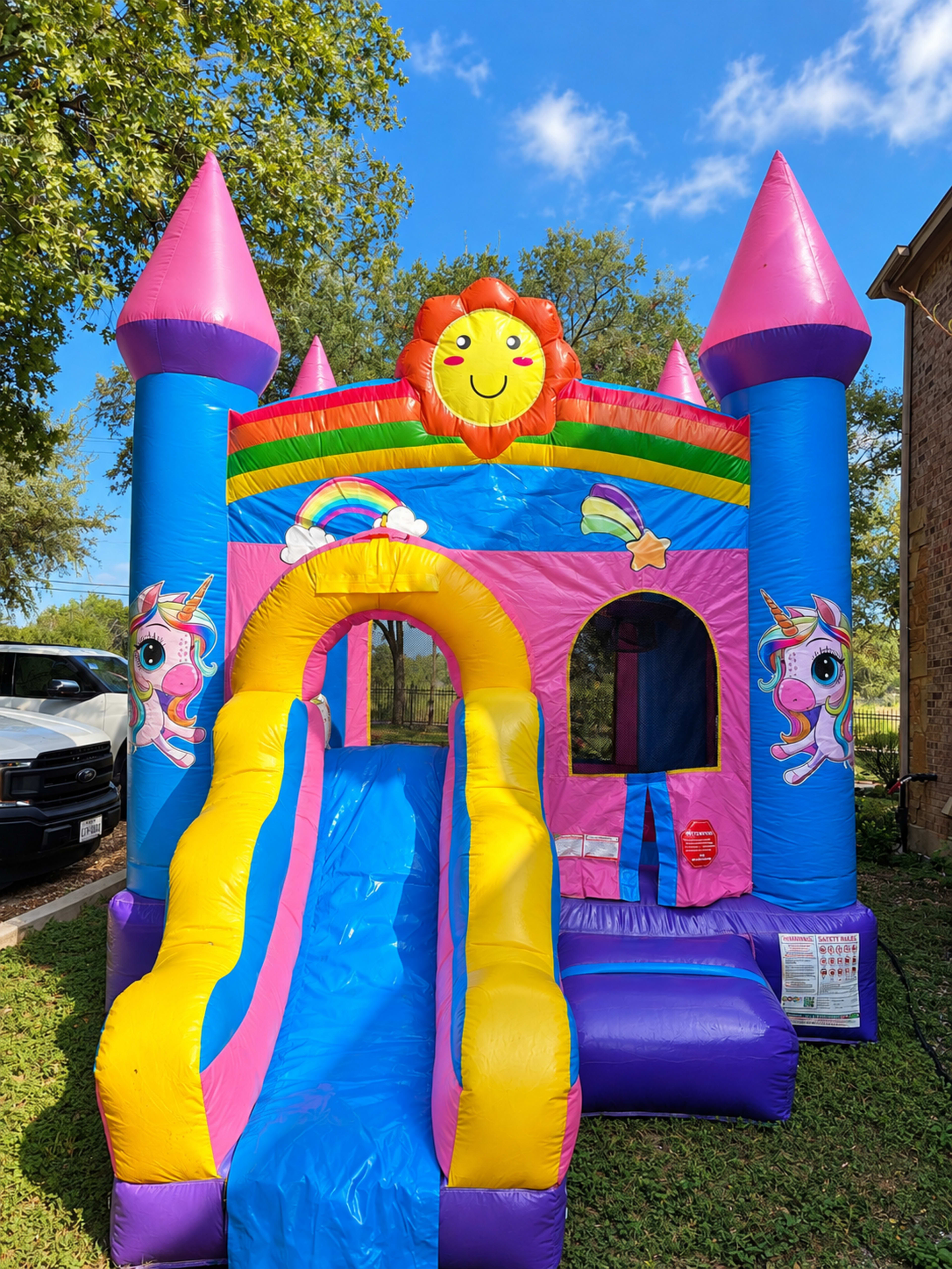 A colorful inflatable bounce house with a slide, featuring a sun and unicorn designs, is set up on a grassy area.