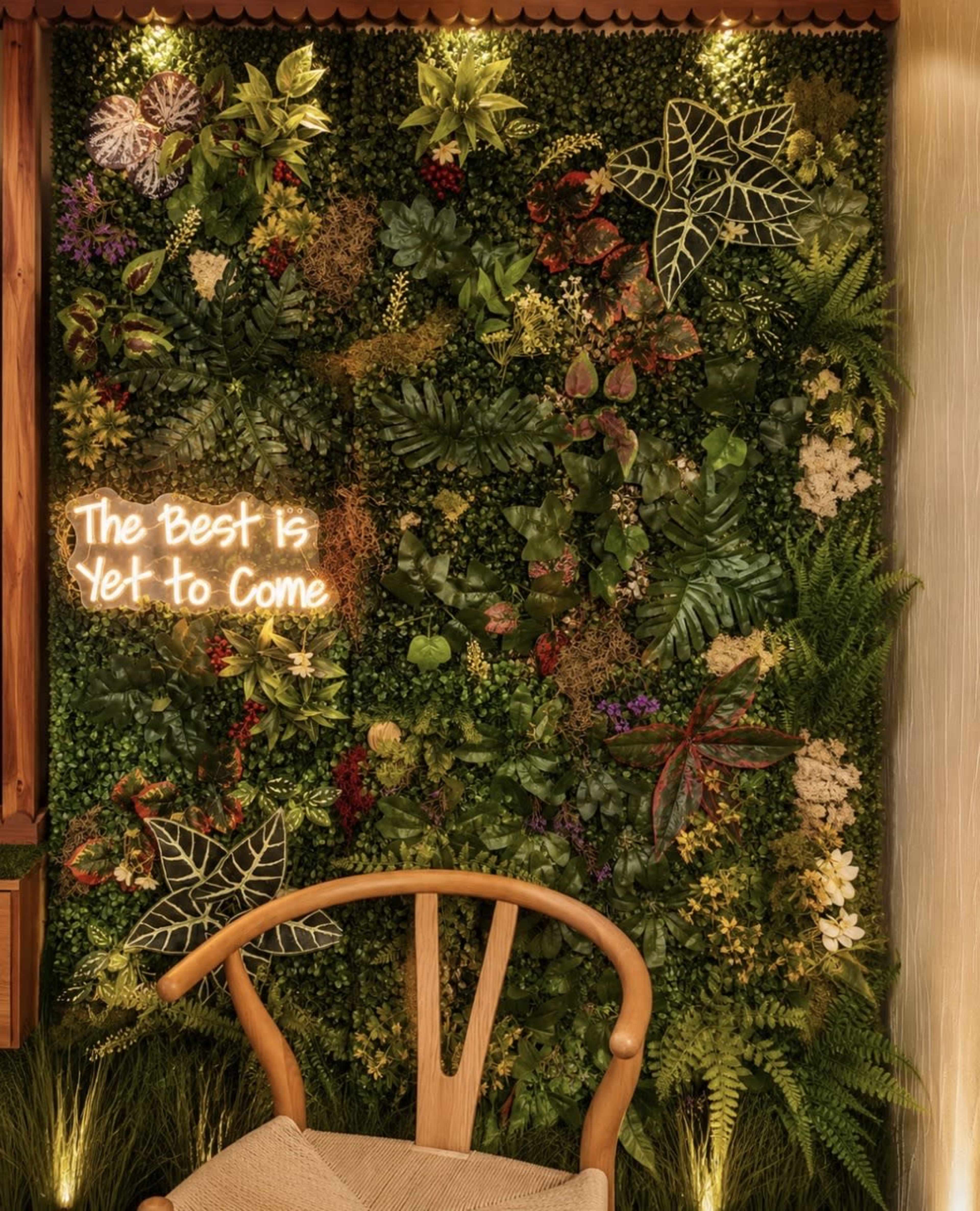 A vibrant green wall adorned with various plants and a glowing neon sign that reads, "The Best is Yet to Come," serves as a backdrop for a wooden chair.