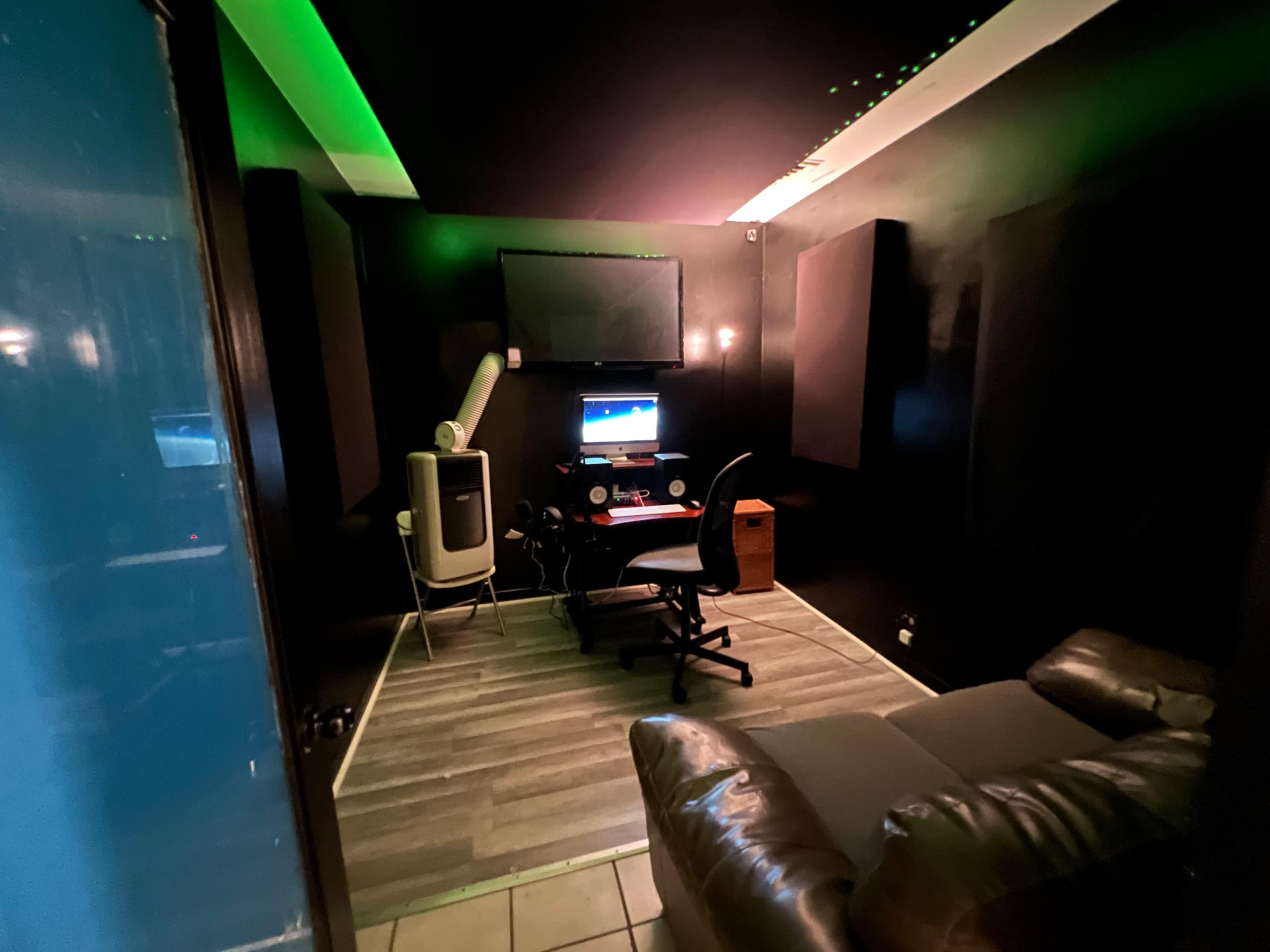 The image shows a dimly lit recording studio with a desk, chair, computer, and acoustic panels on the walls.
