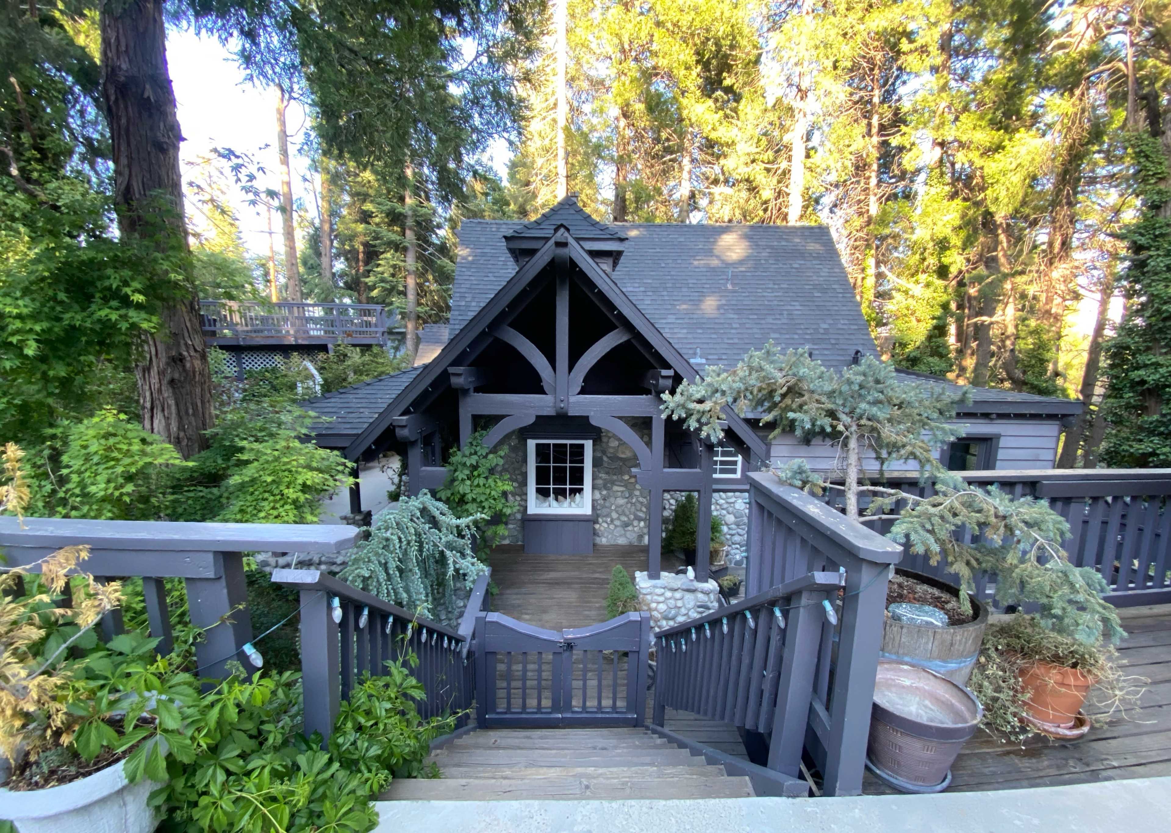 A stone and wood house with a gabled roof is surrounded by tall trees and greenery, featuring a deck and potted plants along the pathway.