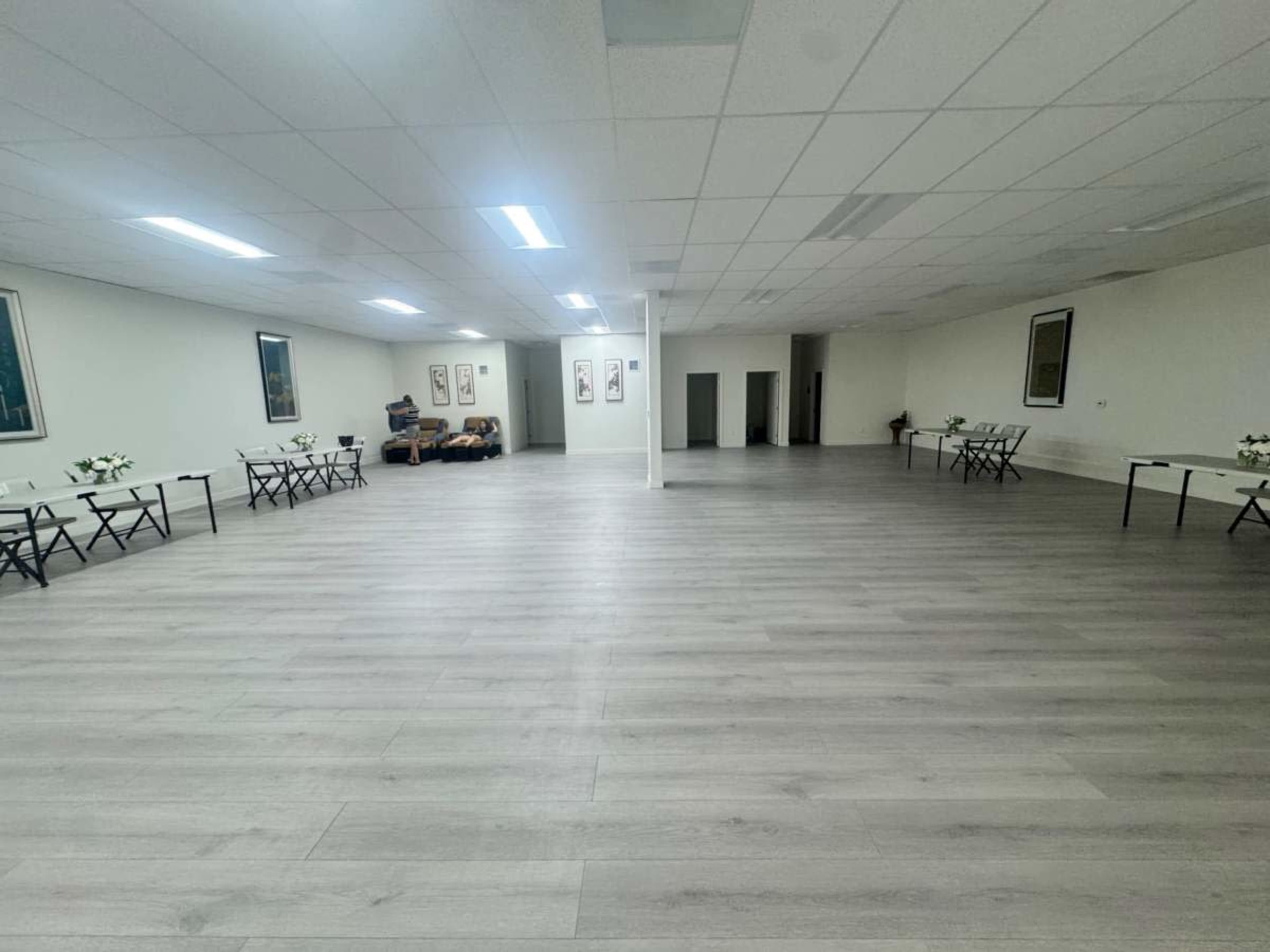 A spacious, well-lit room features empty folding tables arranged along the walls, with a large empty area in the center.