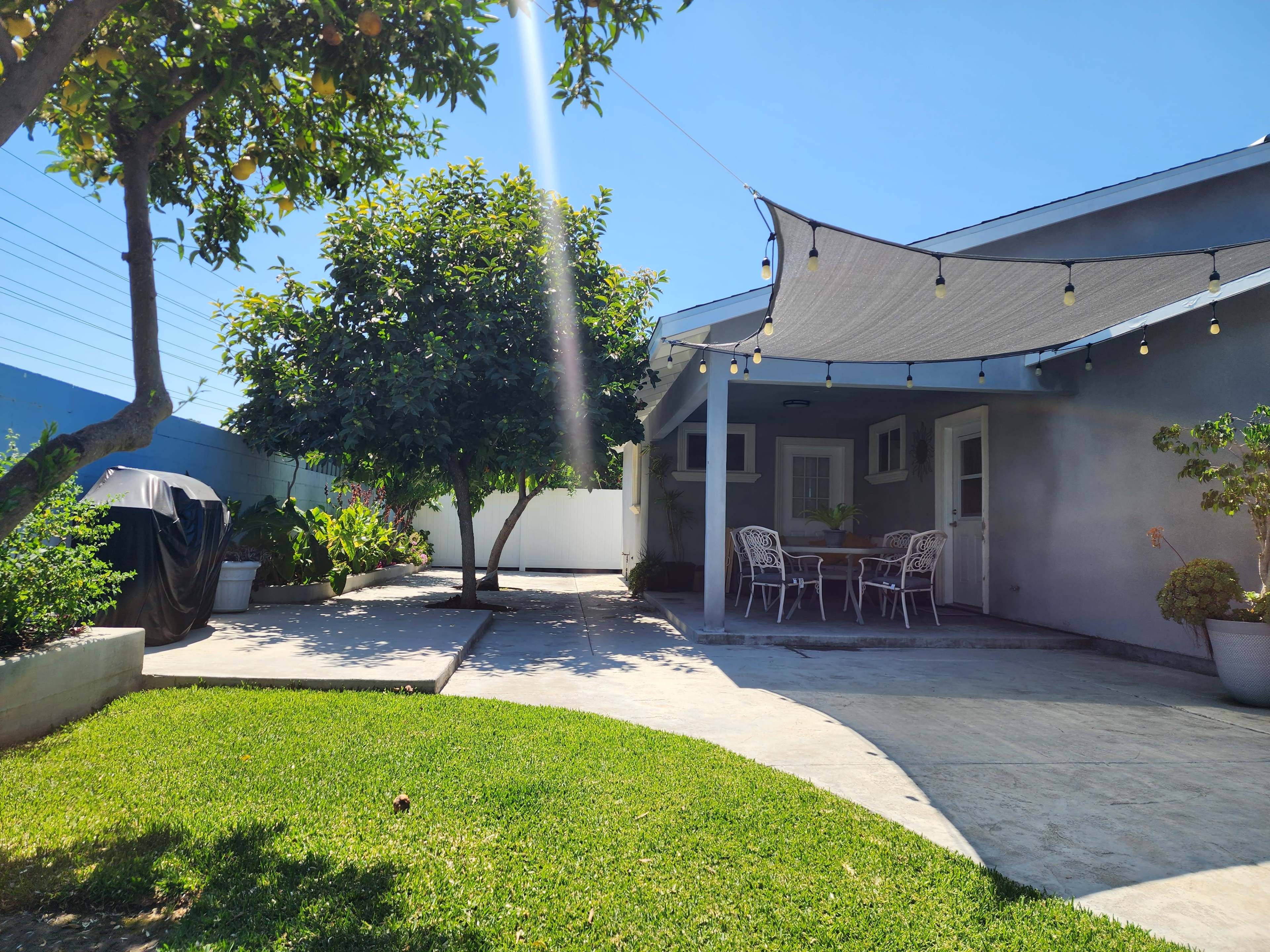 The image shows a backyard with a grassy area, a shaded dining space, a large tree, and a barbecue grill covered with a tarp.
