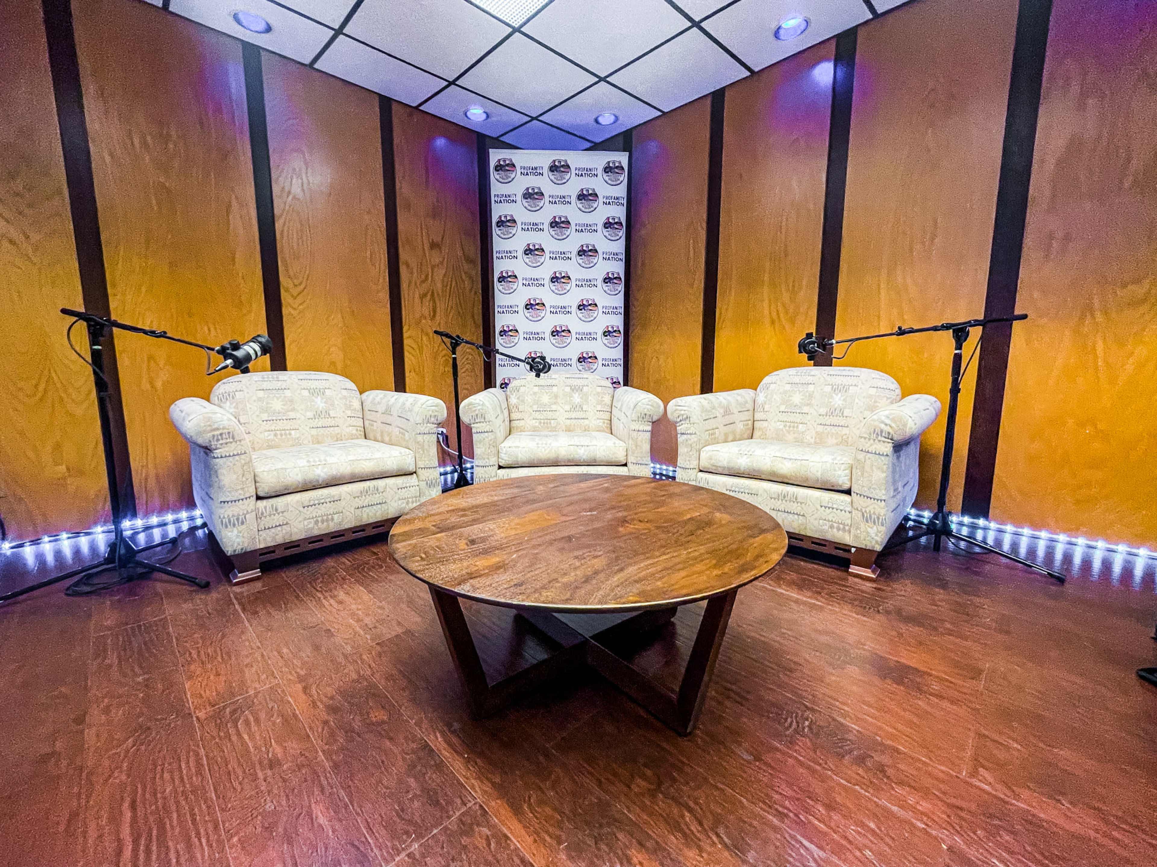 A podcast recording studio featuring two patterned couches, a round wooden table, and microphones positioned on stands.