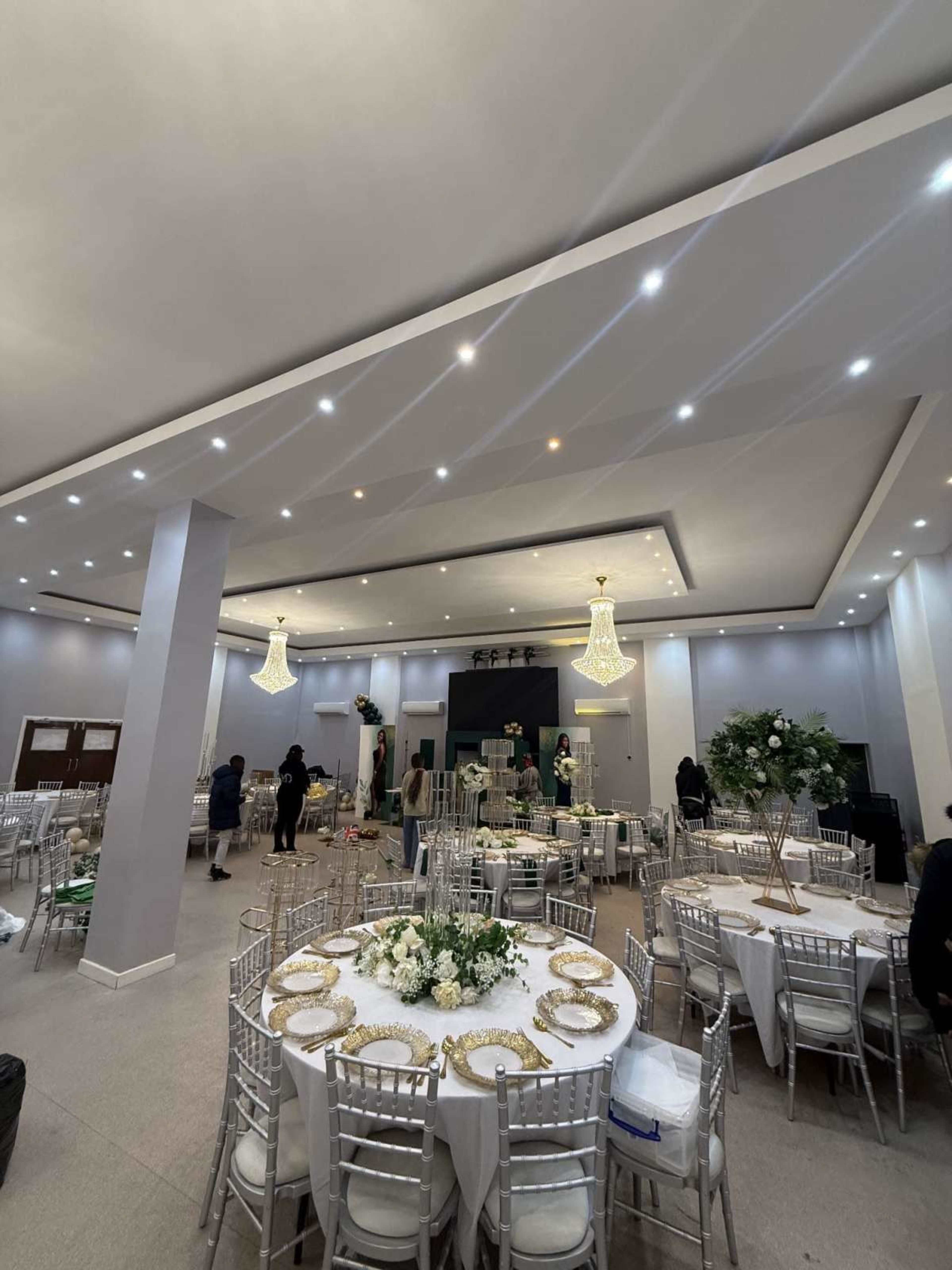 The image shows a banquet hall set up for an event, featuring round tables with white tablecloths and gold accents, surrounded by elegant chandeliers and floral arrangements.