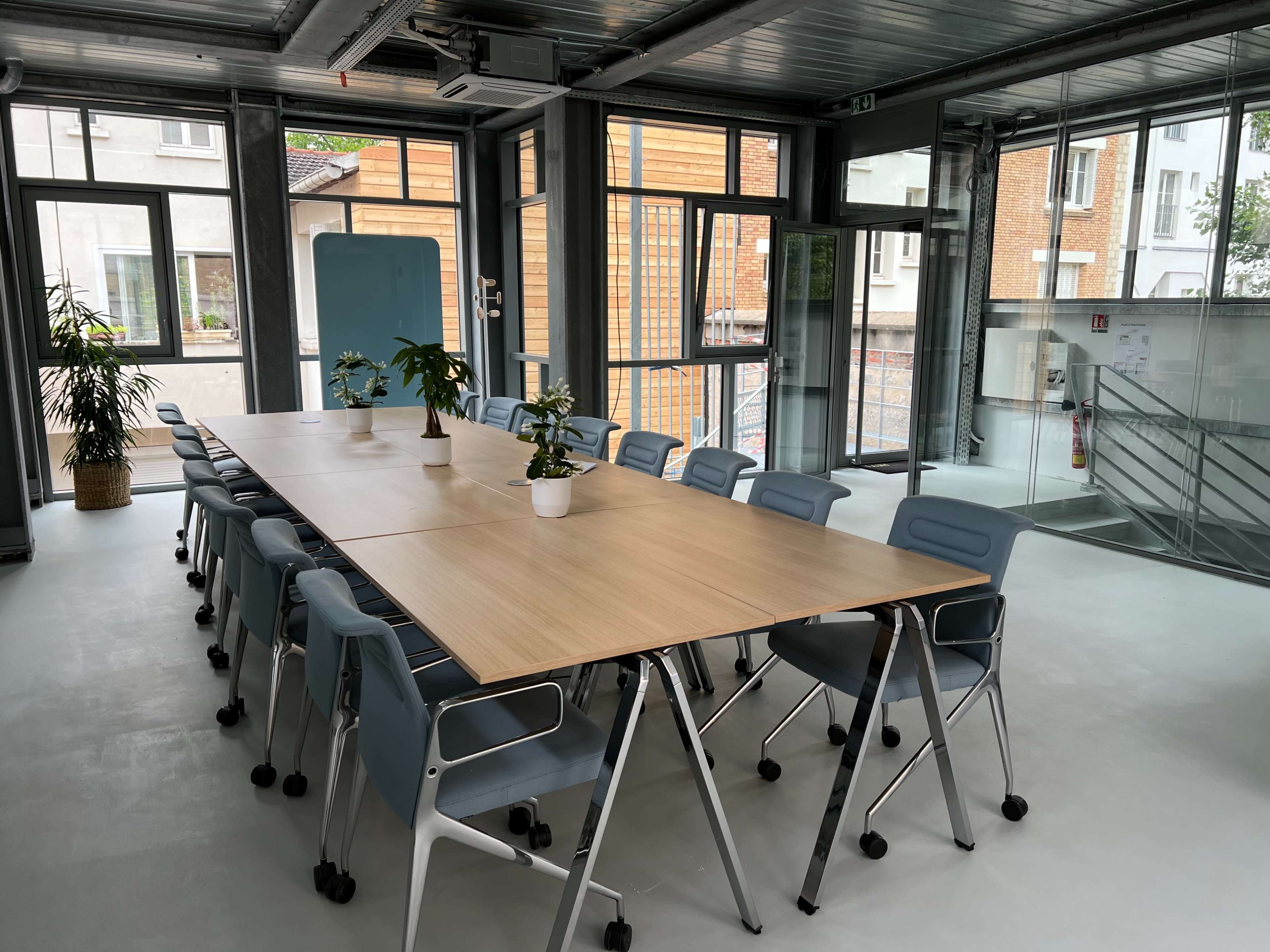 A modern conference room with a long wooden table surrounded by blue office chairs and large windows allowing natural light.