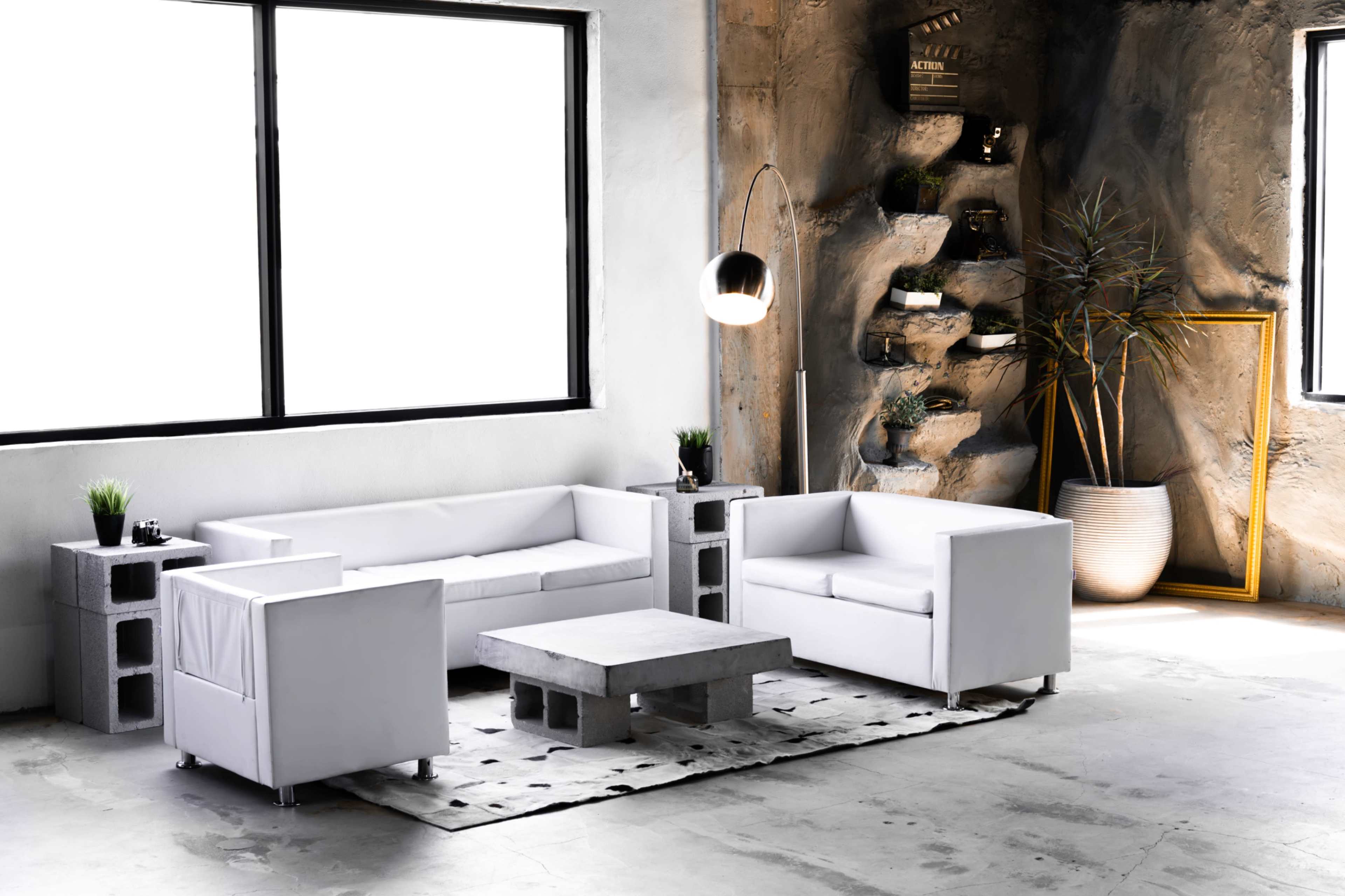 A modern living room features white sofas arranged around a concrete coffee table on a patterned rug, with large windows and a textured wall adorned with plants and decorations.