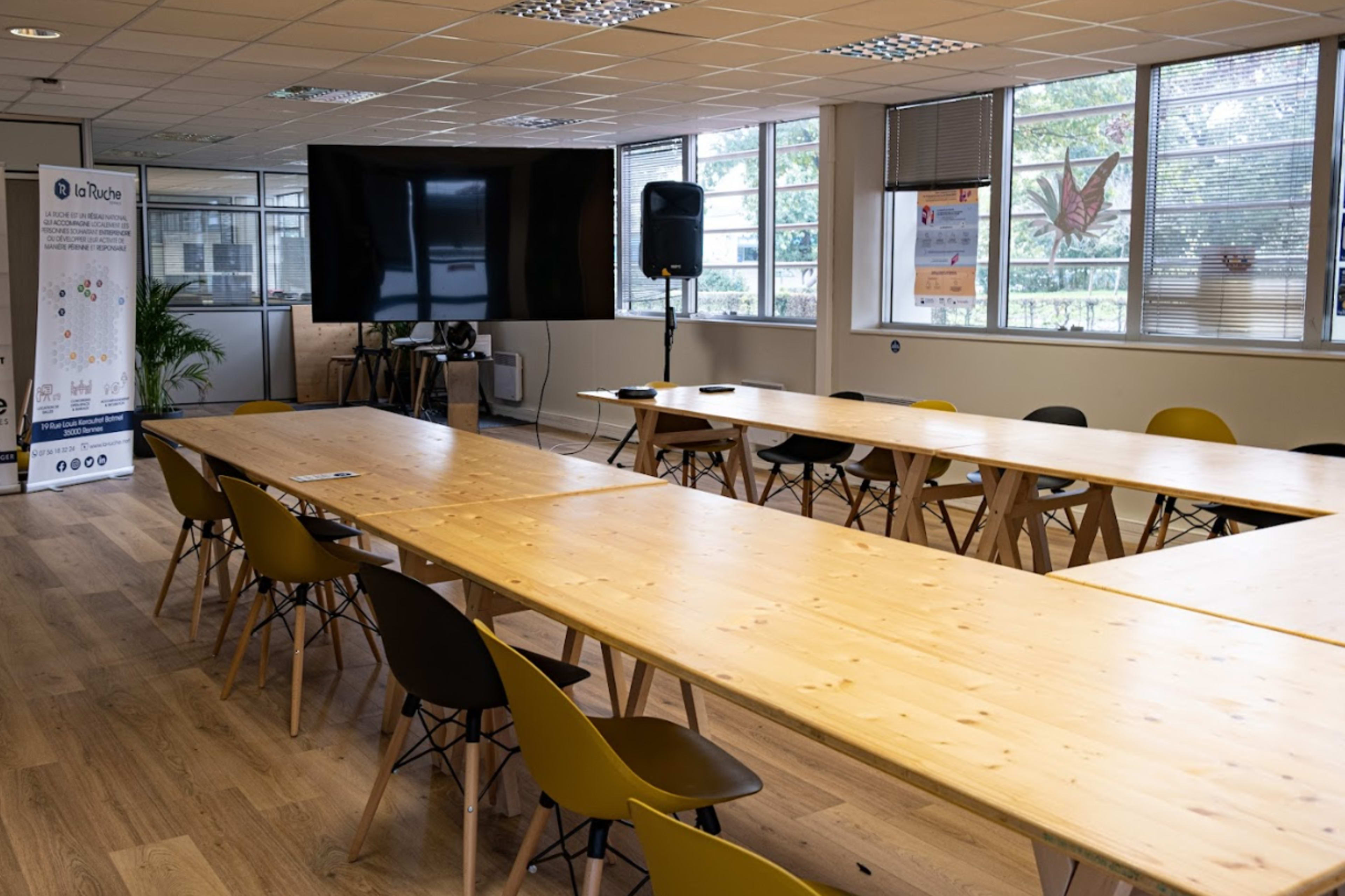 A spacious meeting room features long wooden tables arranged in a U-shape, surrounded by chairs, with large windows allowing natural light and a screen for presentations.