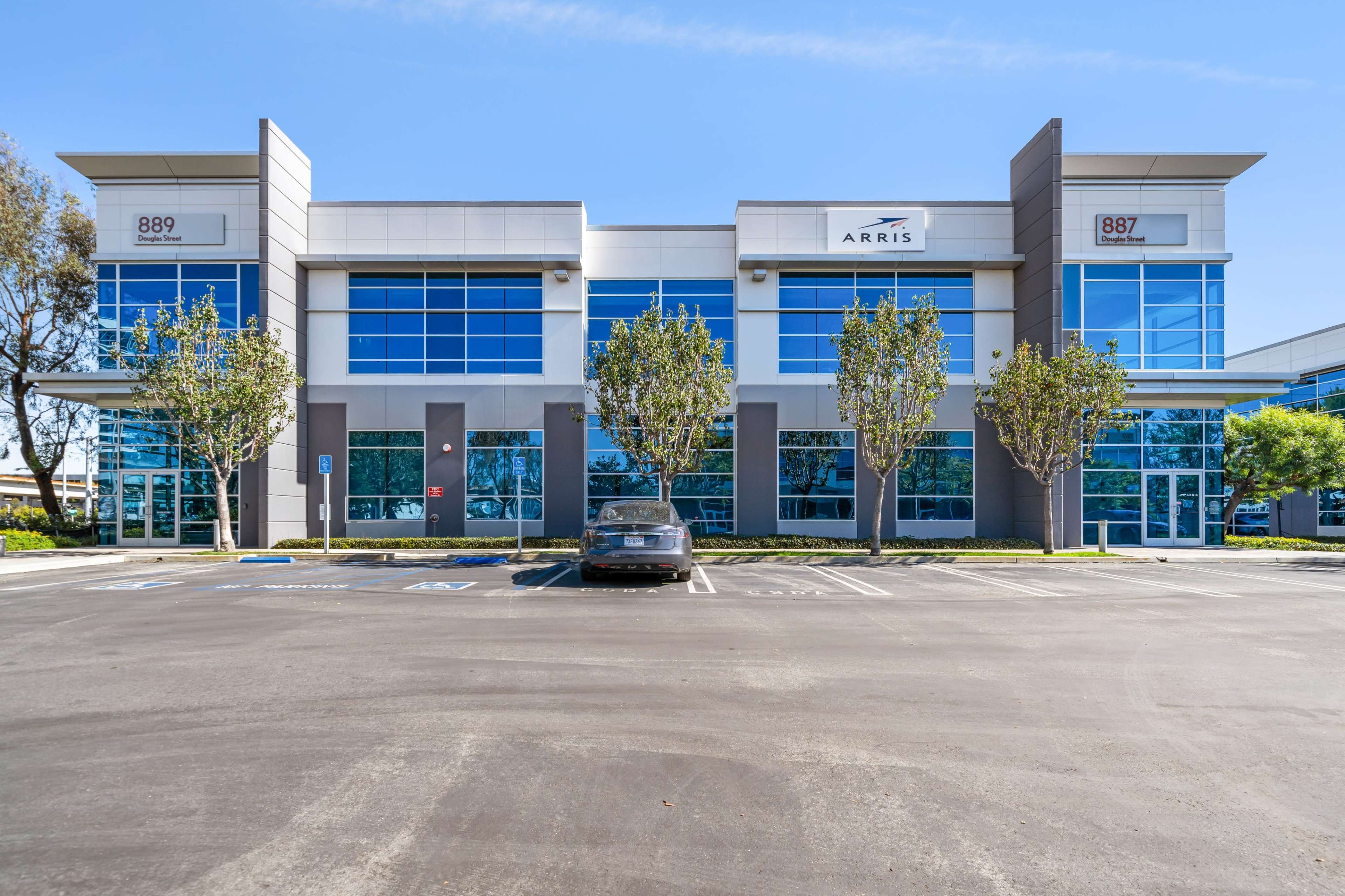 A modern office building with large glass windows and two prominent entrance signs, located in a parking lot with a black car parked in front.