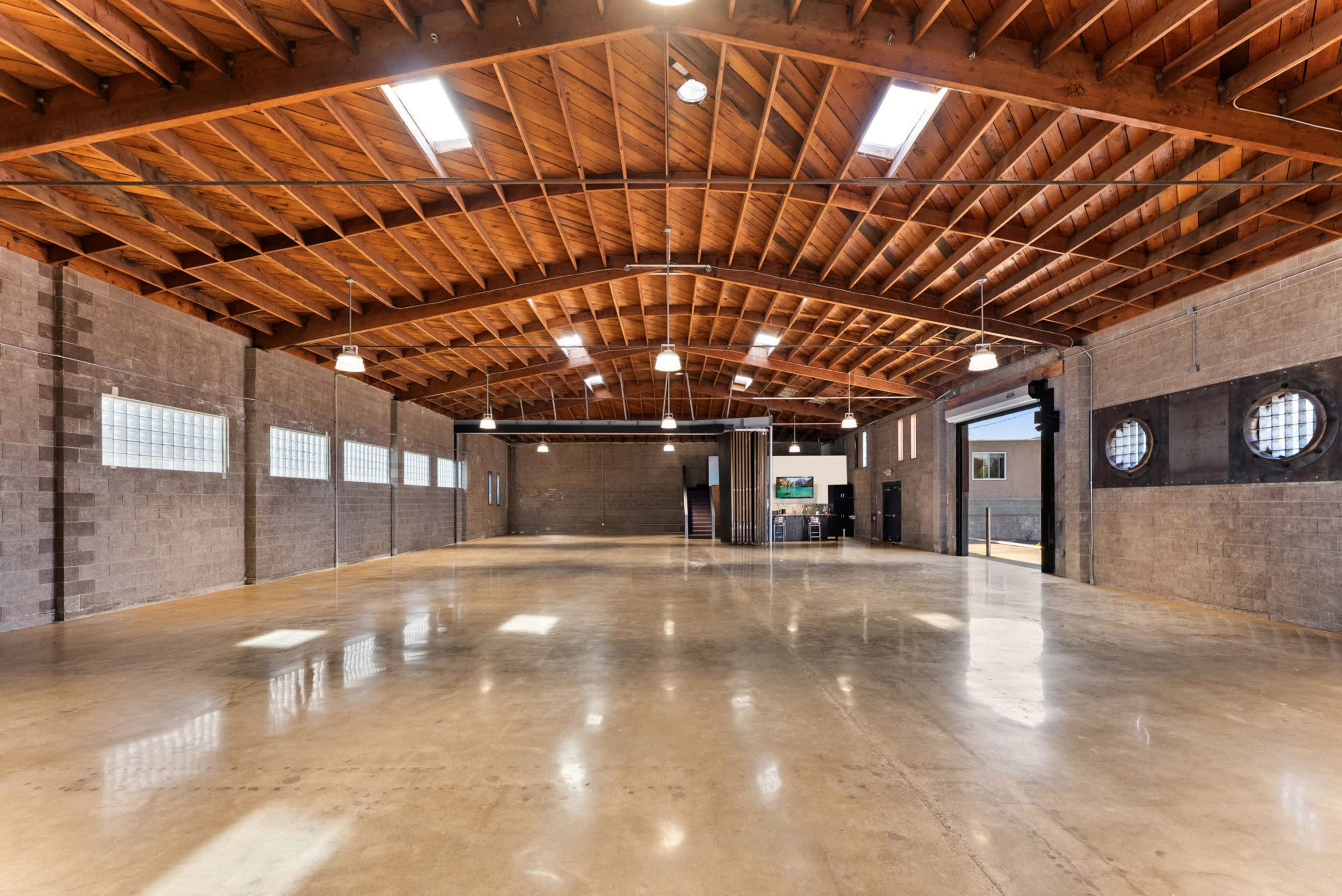 The image shows a spacious industrial-style interior with high wooden ceilings, concrete flooring, and large windows along the walls.