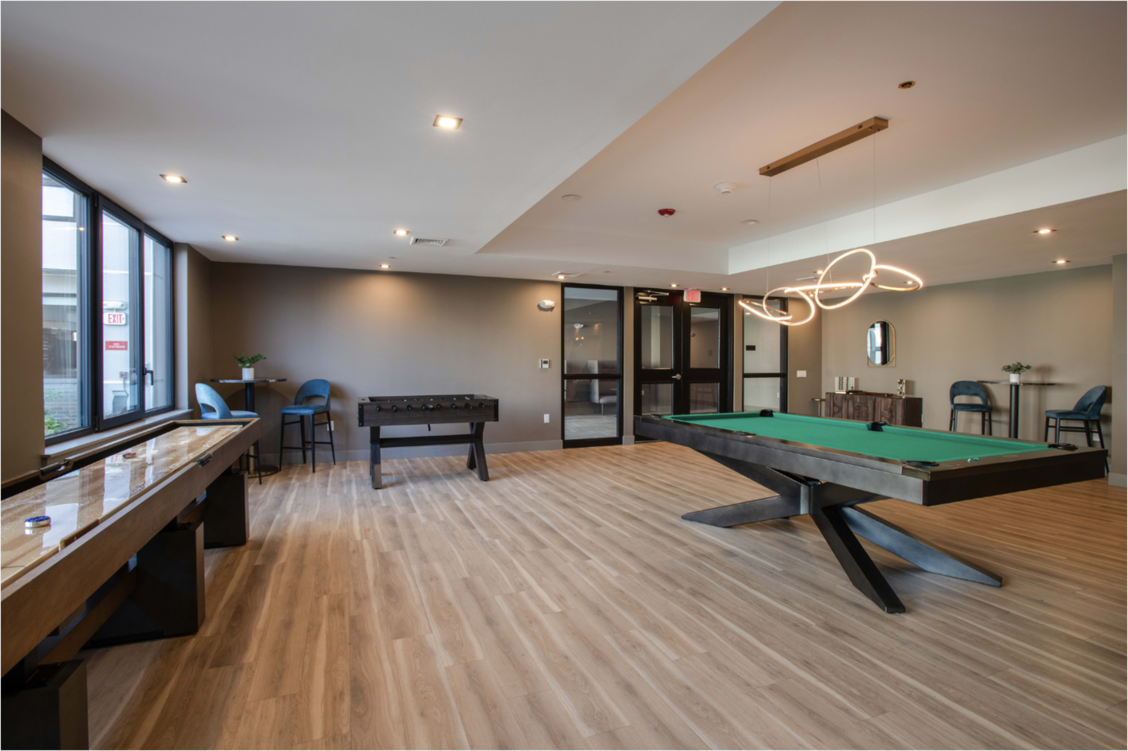The image shows a modern game room with a green-felt pool table, foosball table, and seating areas, illuminated by contemporary lighting fixtures.