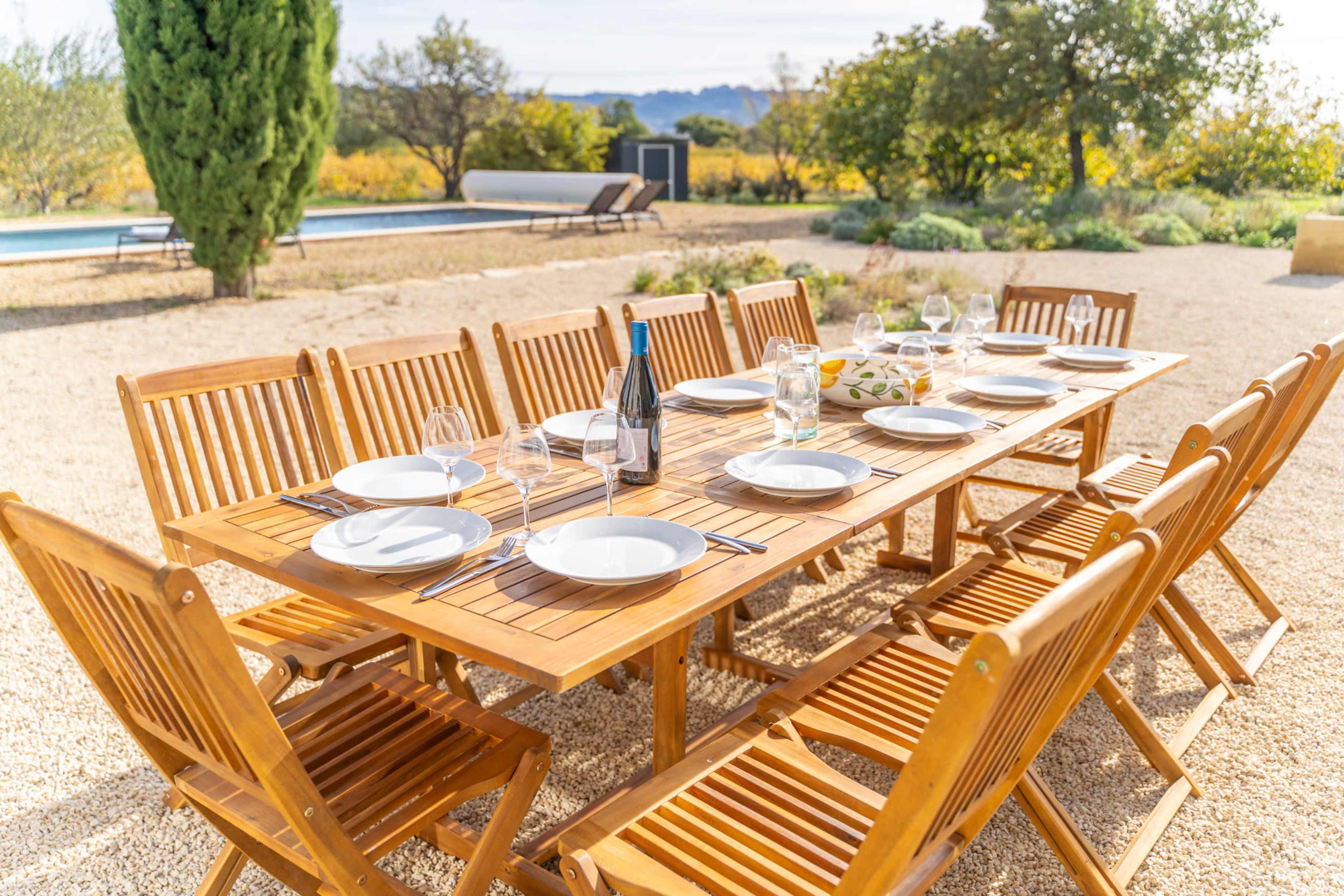 A wooden dining table is set outdoors with plates, glasses, and a bottle, surrounded by chairs in a vineyard landscape.