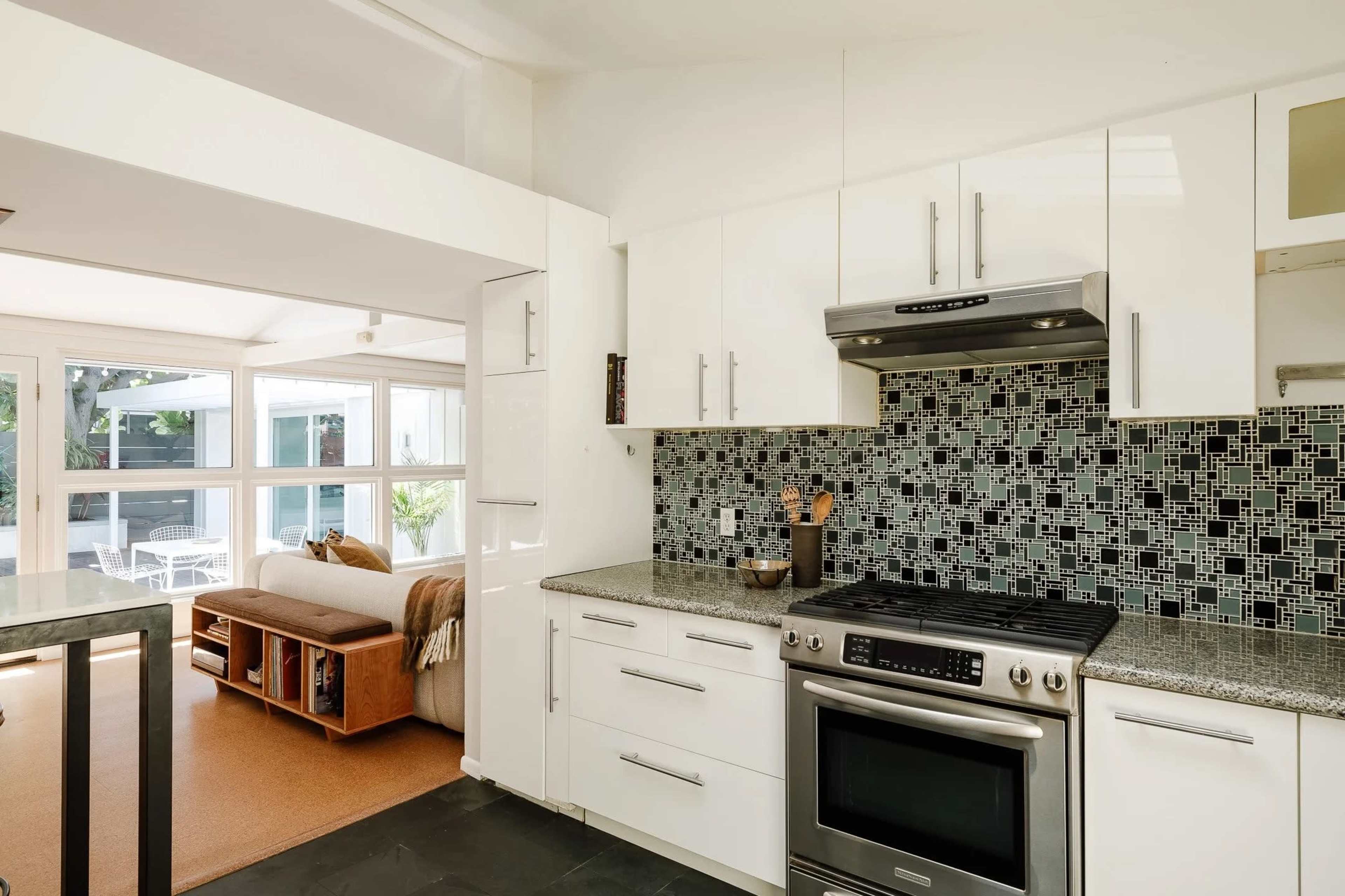 The image shows a modern kitchen featuring white cabinets, stainless steel appliances, and a mosaic tile backsplash complemented by a spacious open layout leading to a living area.