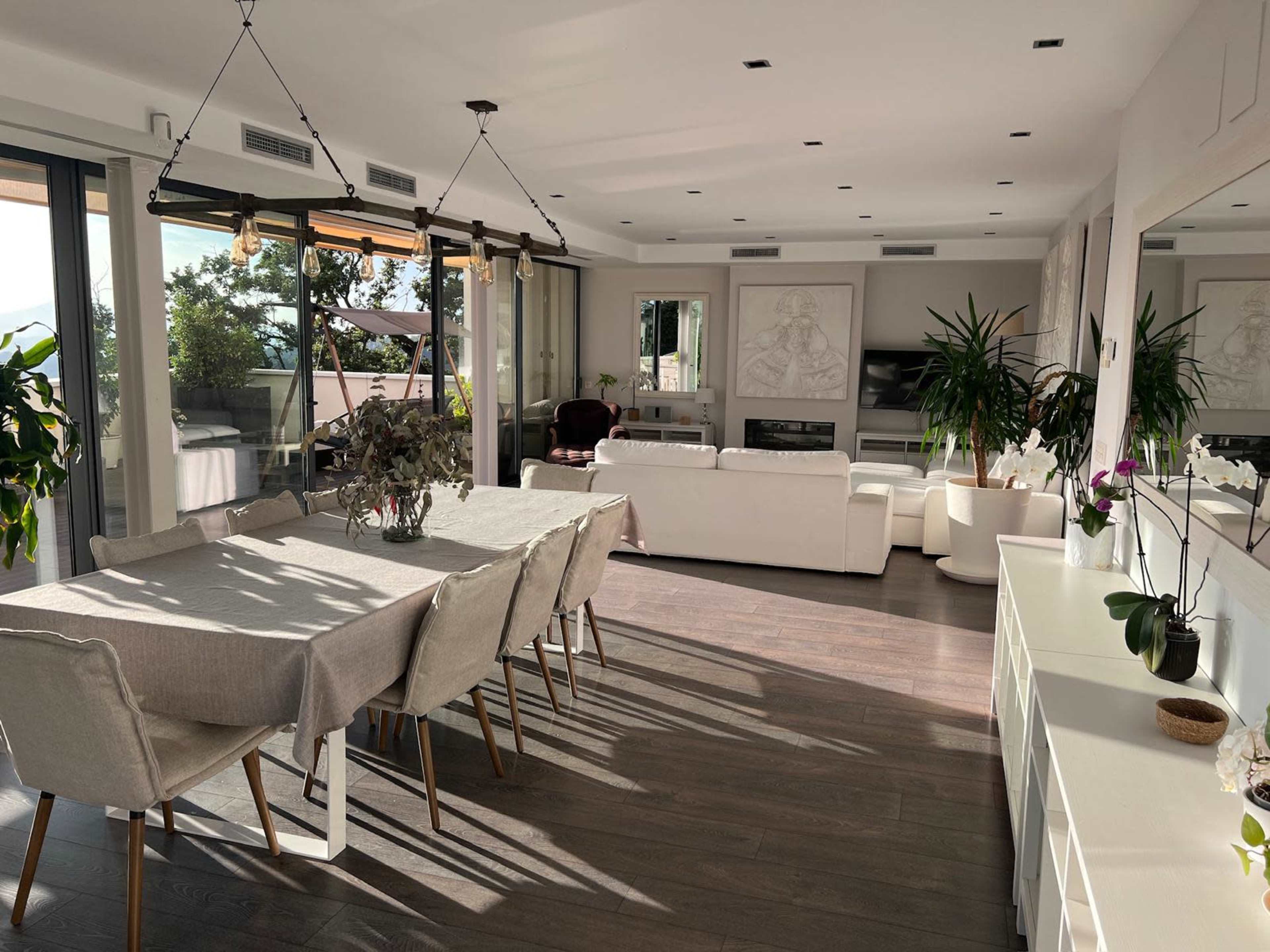 The image shows a modern, open-concept living and dining area featuring a long dining table, a white sofa, and large windows that allow natural light to enter.