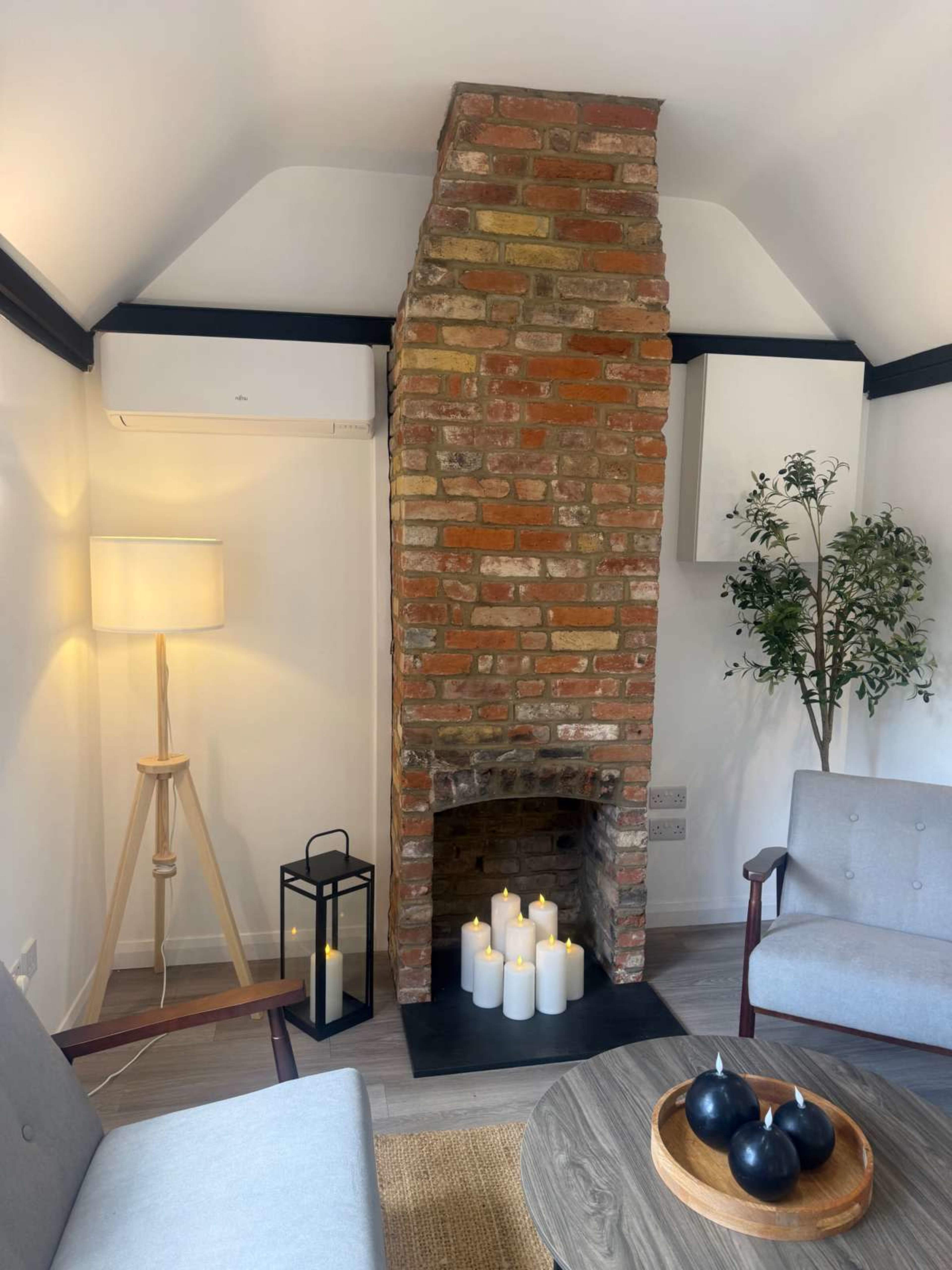 The image shows a cozy living space featuring a brick fireplace, a floor lamp, a candle arrangement, and two armchairs.