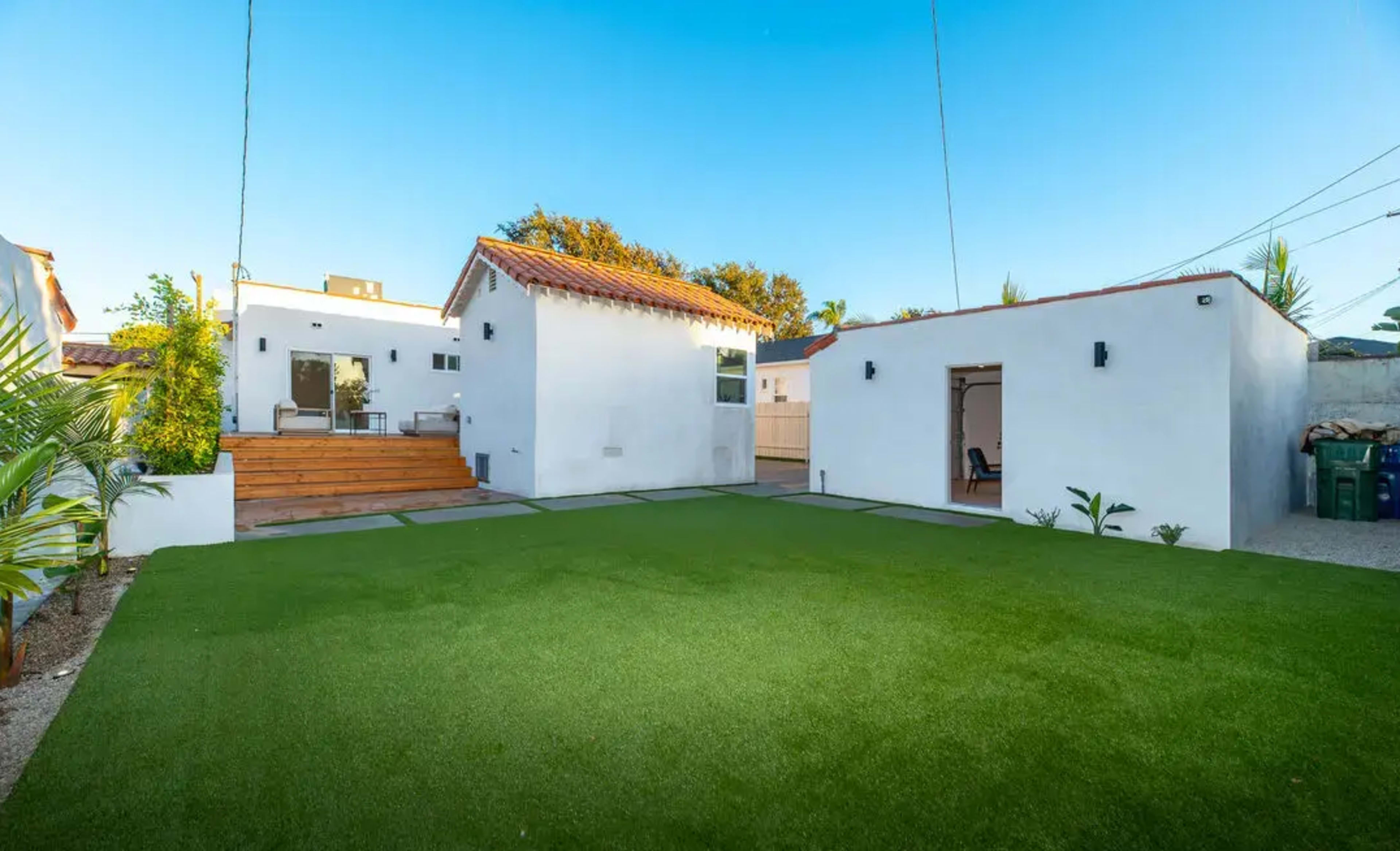 The image shows a modern backyard with two white buildings, a green artificial lawn, and a wooden deck area.