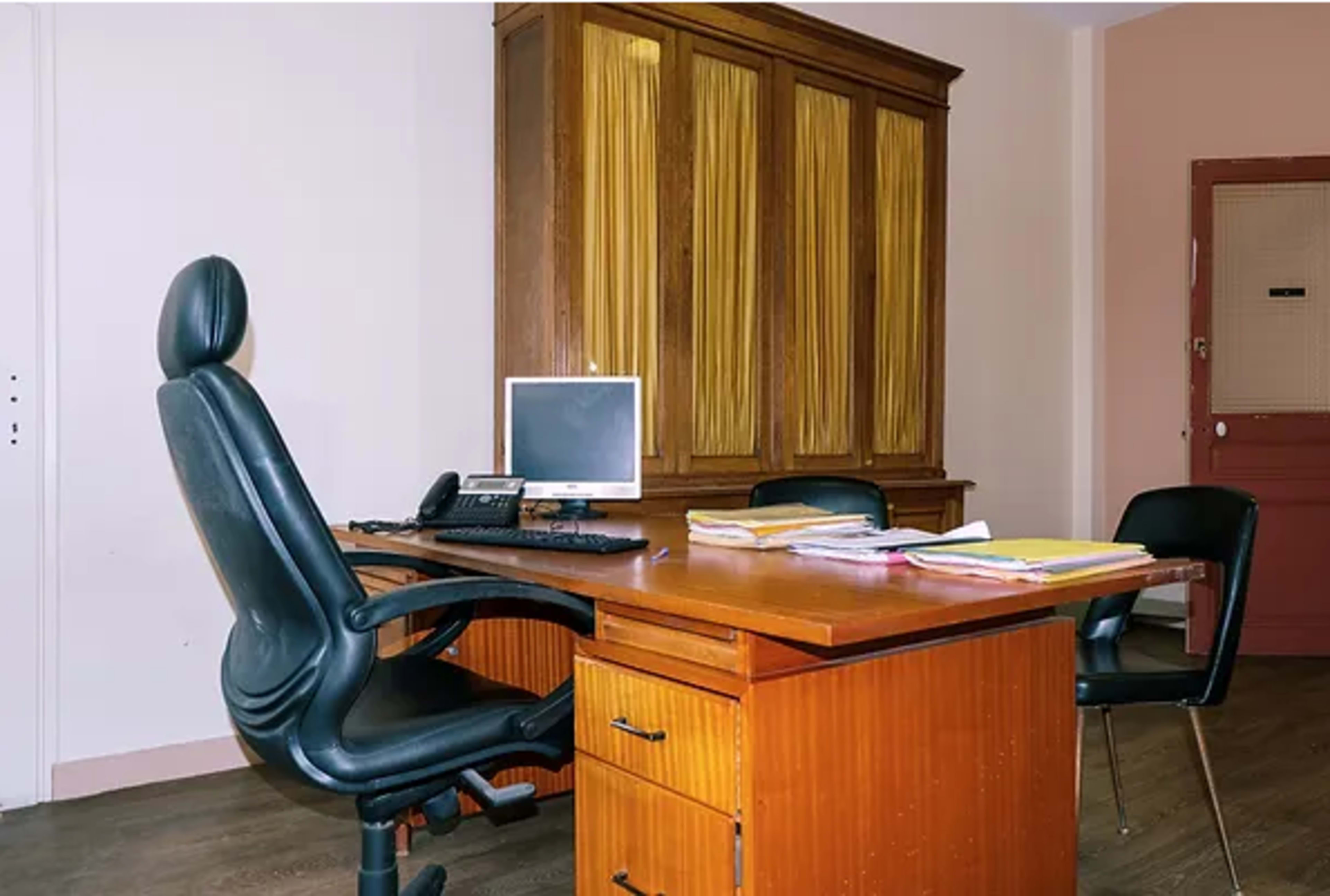 An office space featuring a wooden desk, a swivel chair, a computer monitor, and stack of papers.