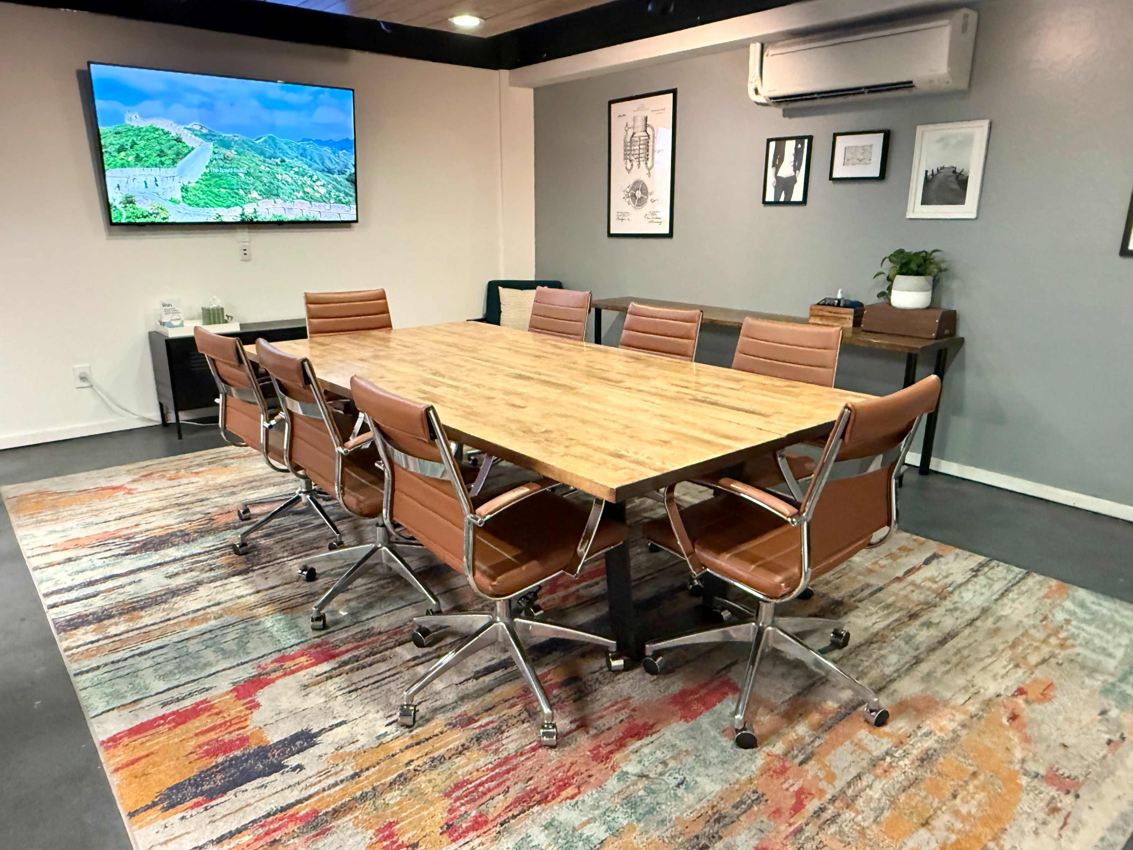 The image shows a modern conference room with a large wooden table surrounded by leather chairs, a television on the wall, and decorative artwork.