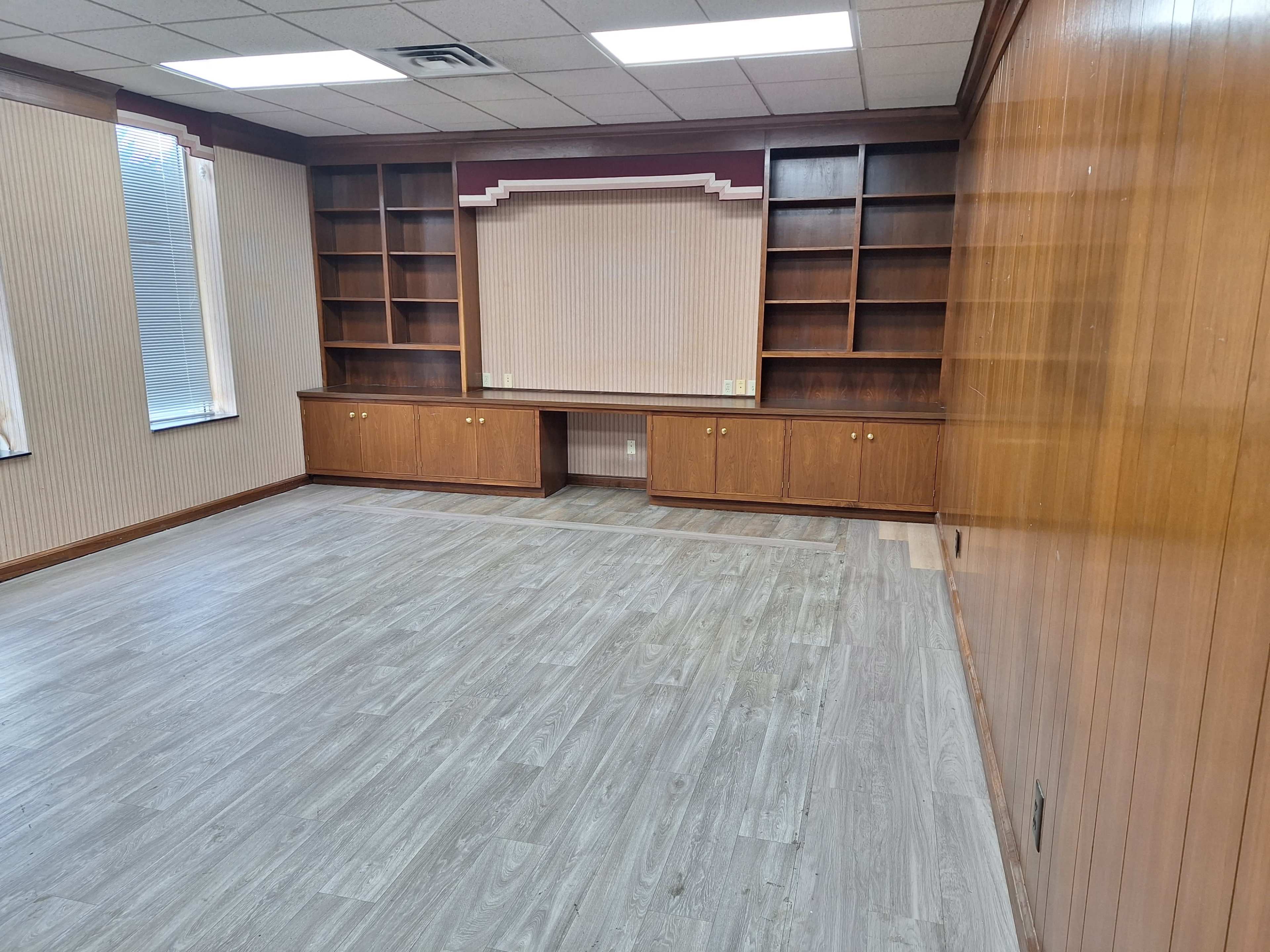 The image shows a sparsely furnished room with wooden paneling, a built-in shelf unit, and large windows providing natural light.