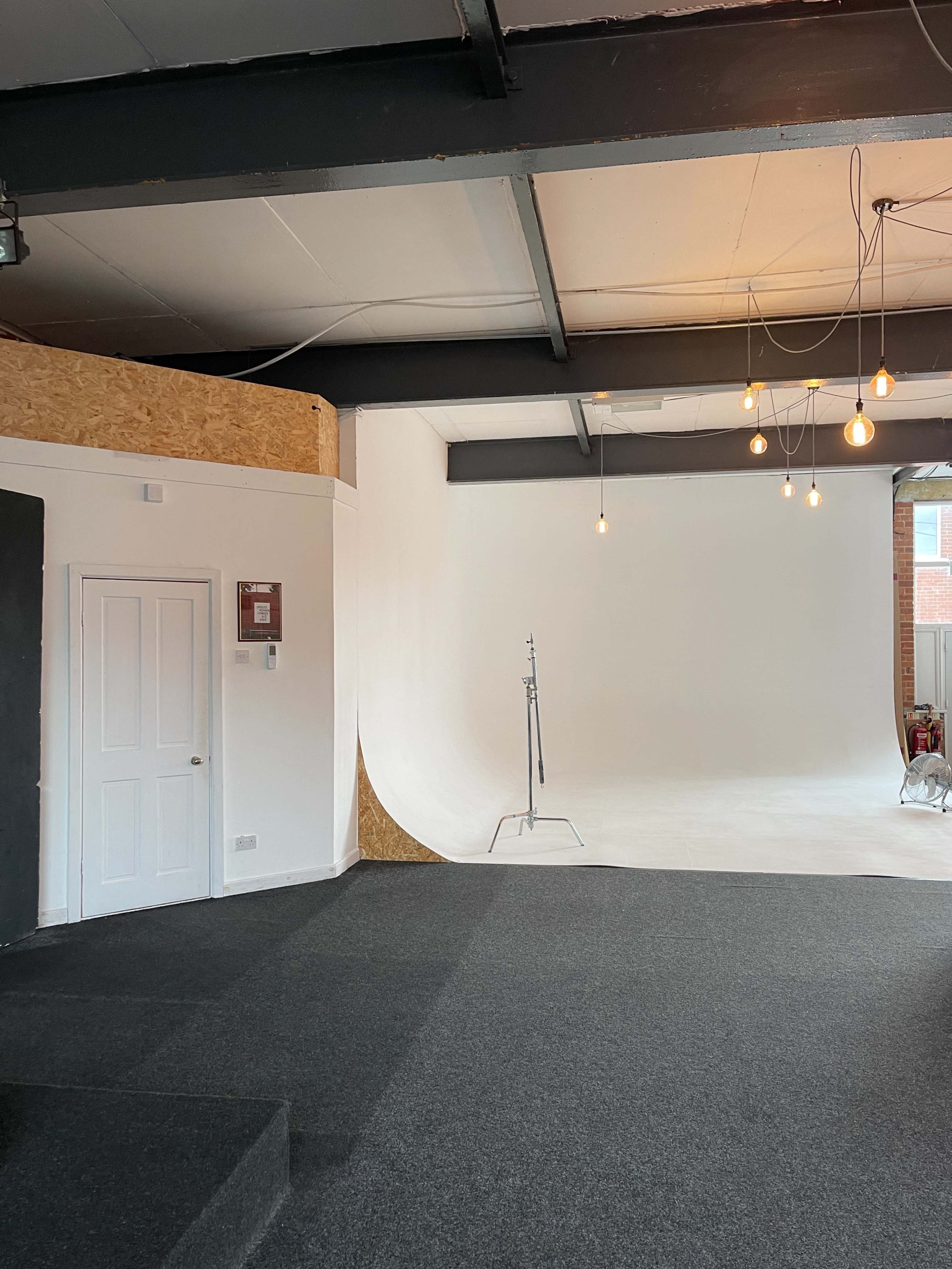 A spacious studio with a seamless white backdrop, a clothing rack, and hanging light bulbs, featuring gray carpeted flooring and a door on the left.