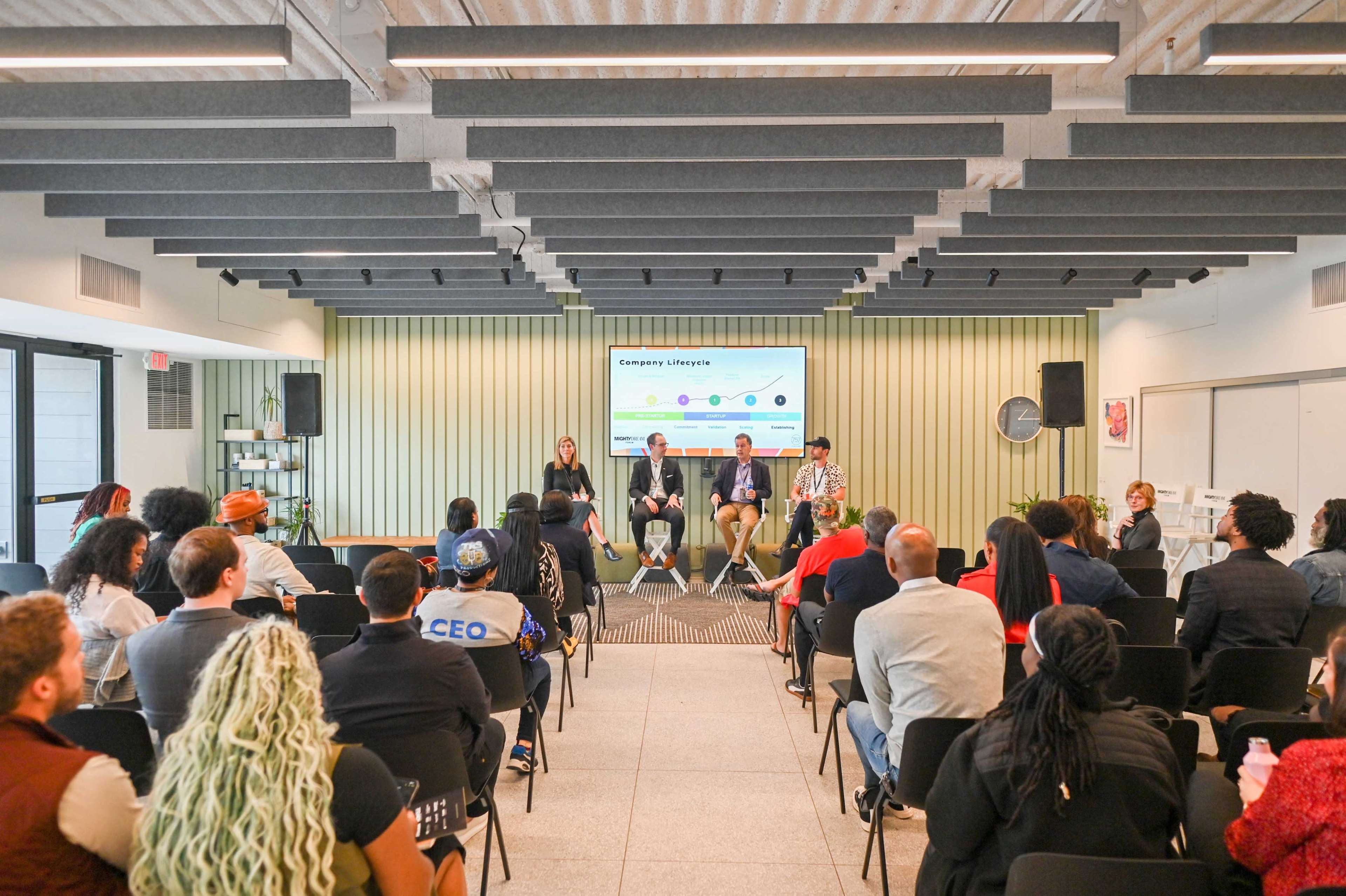 A panel discussion is taking place in a modern conference room with attendees seated in rows facing the speakers.