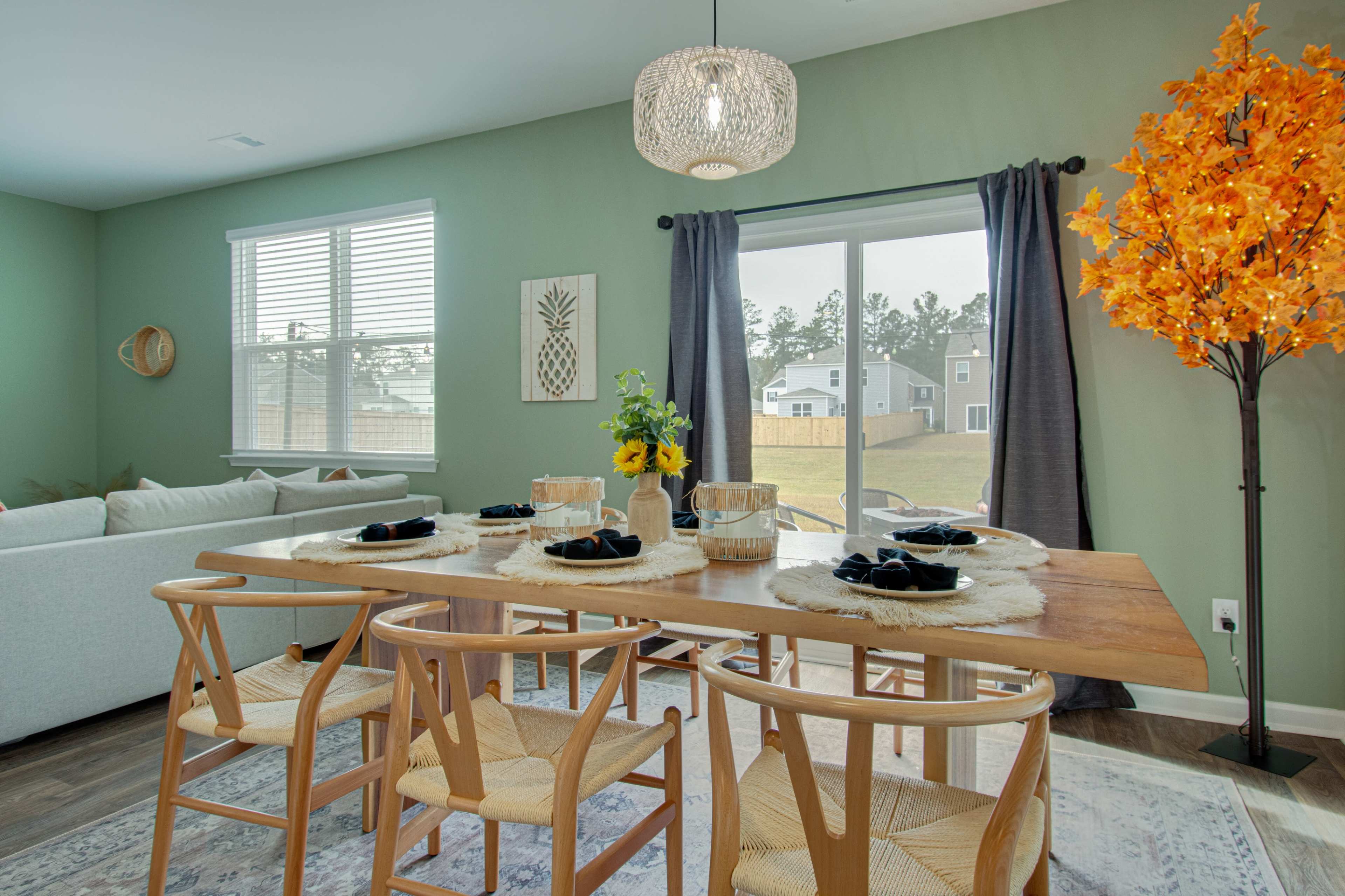A modern dining area features a wooden table set for six, accompanied by light-colored chairs, a vibrant artificial tree, and large windows overlooking a backyard.