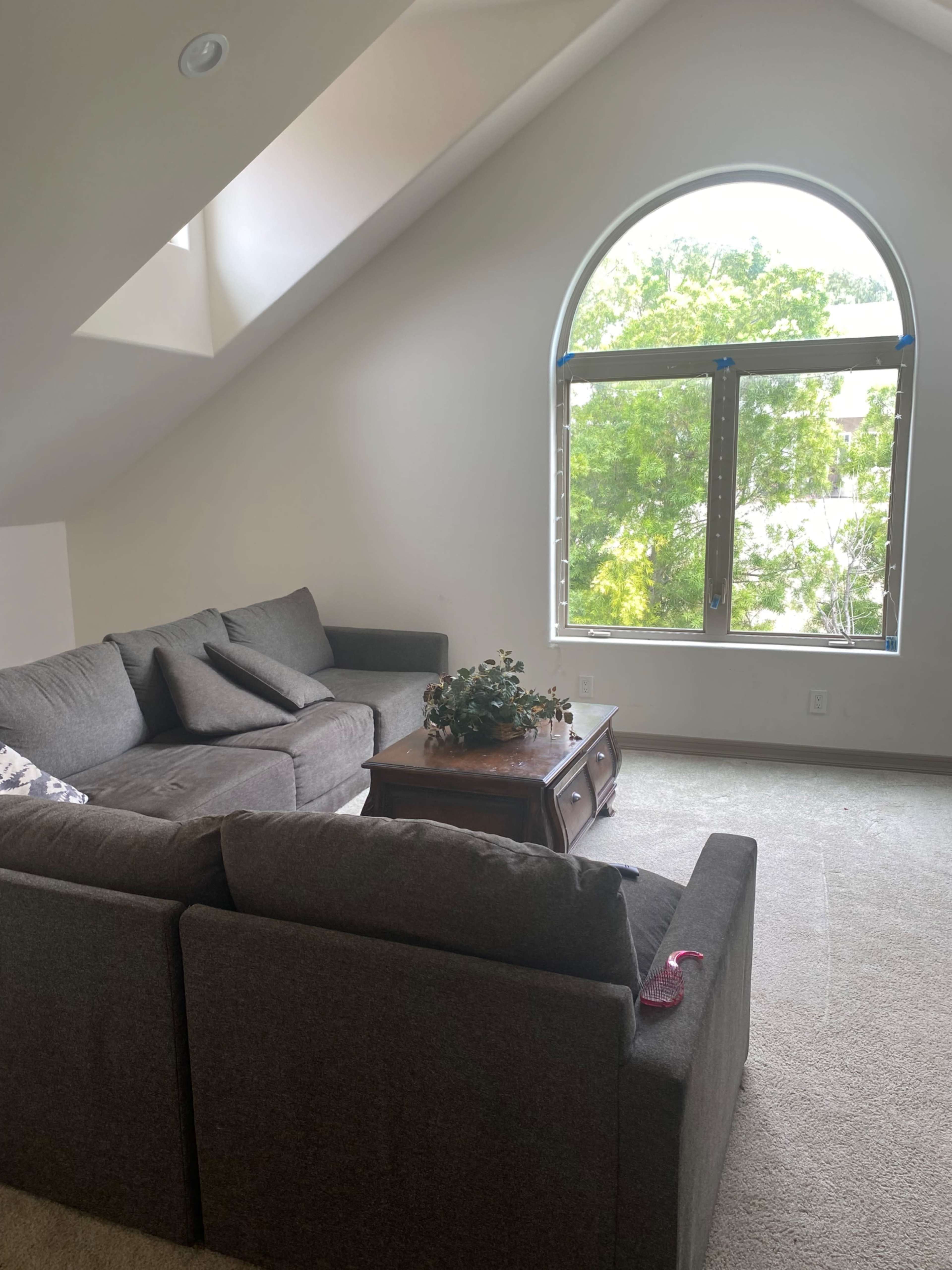 The image shows a gray sectional sofa arranged around a wooden coffee table in a room with a large arched window that provides a view of greenery outside.