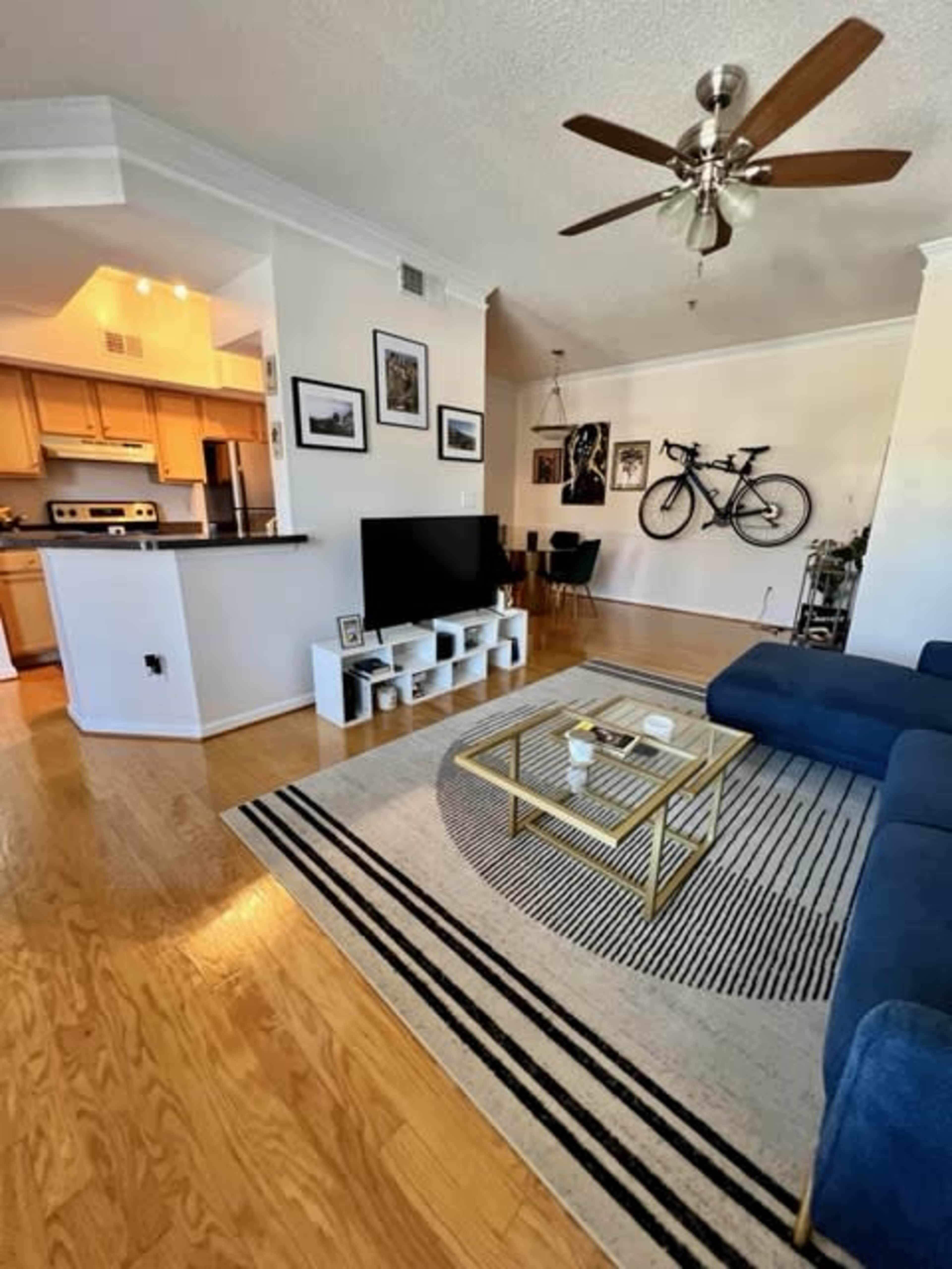 A spacious living room features a blue sectional sofa, a coffee table, and a decorative rug, with a kitchen and dining area visible in the background.