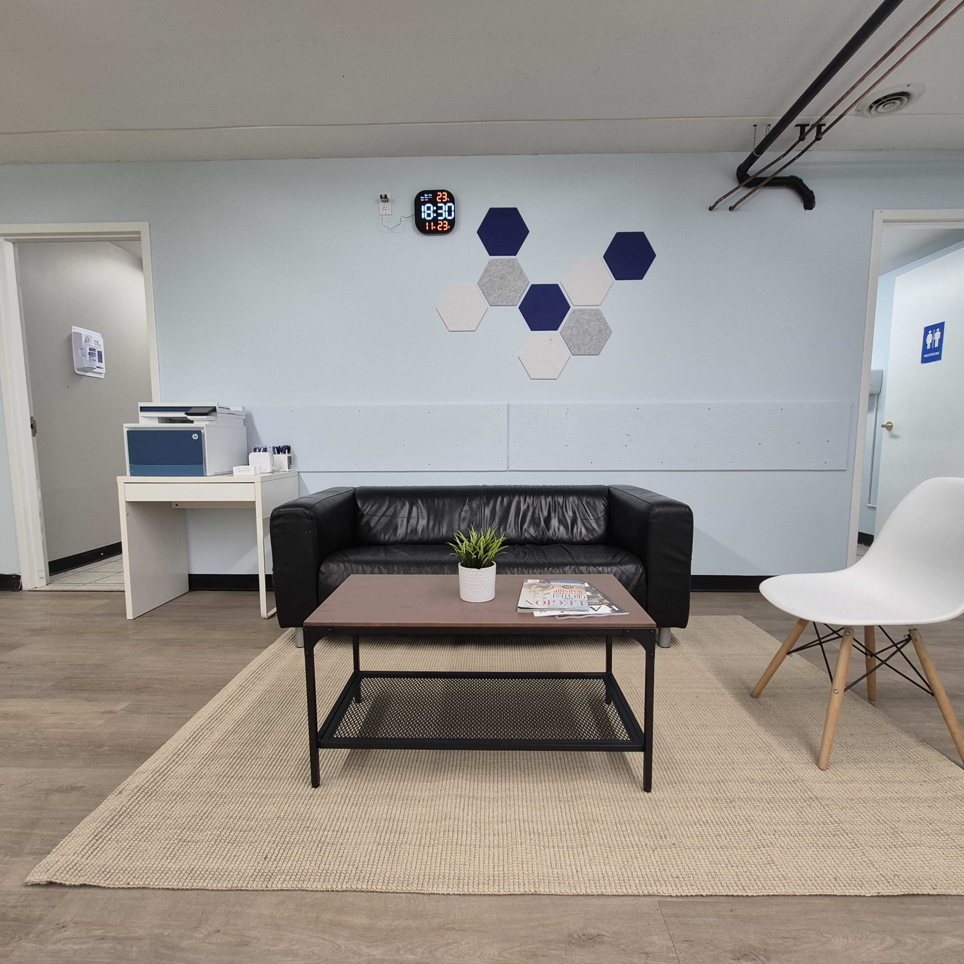 The image shows a waiting area with a black couch, a coffee table, a white chair, and a small plant on the table, all set against light blue walls and a rug.