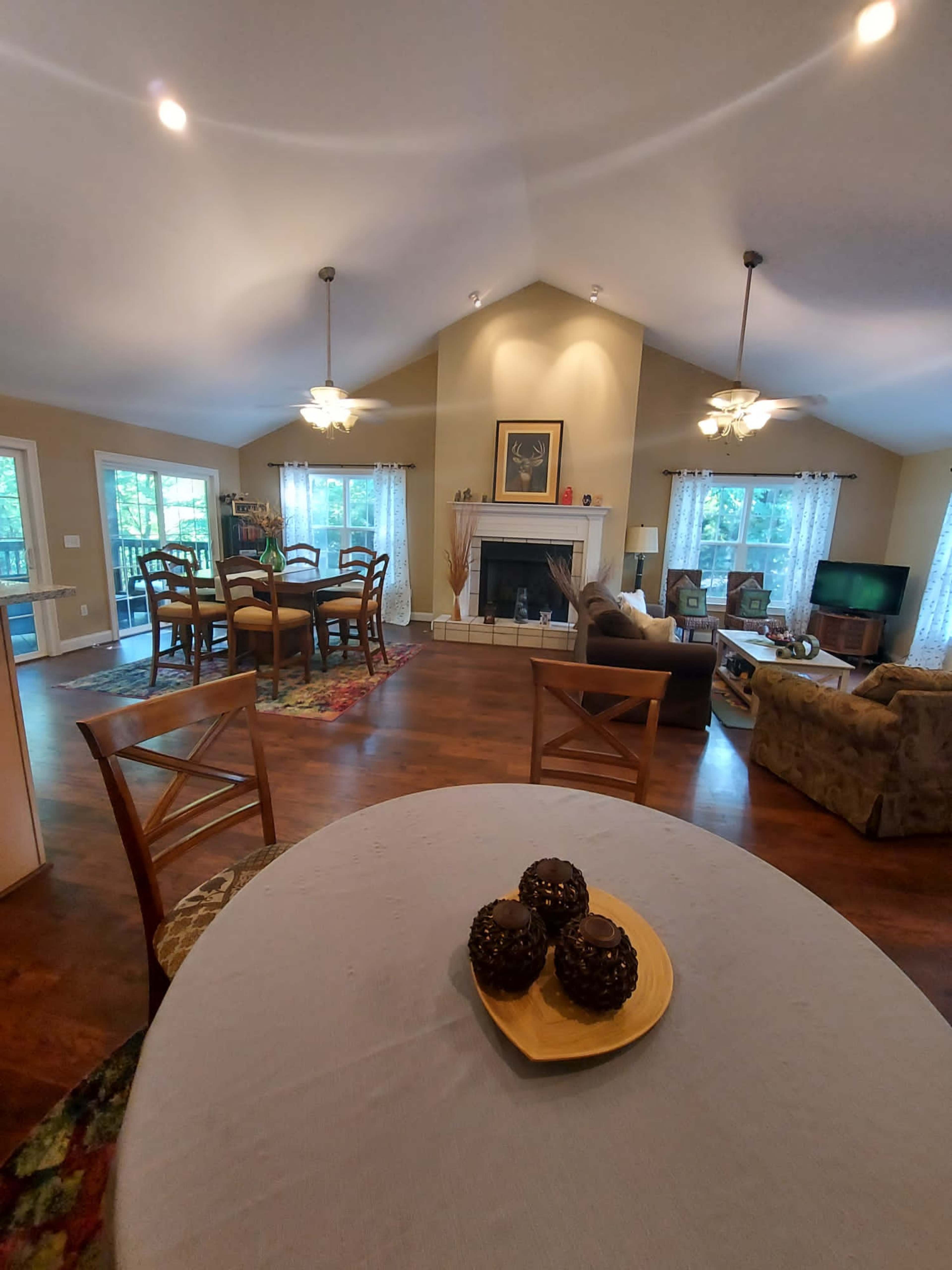 A spacious living area features a dining table with decorative items, a fireplace surrounded by seating, and large windows letting in natural light.