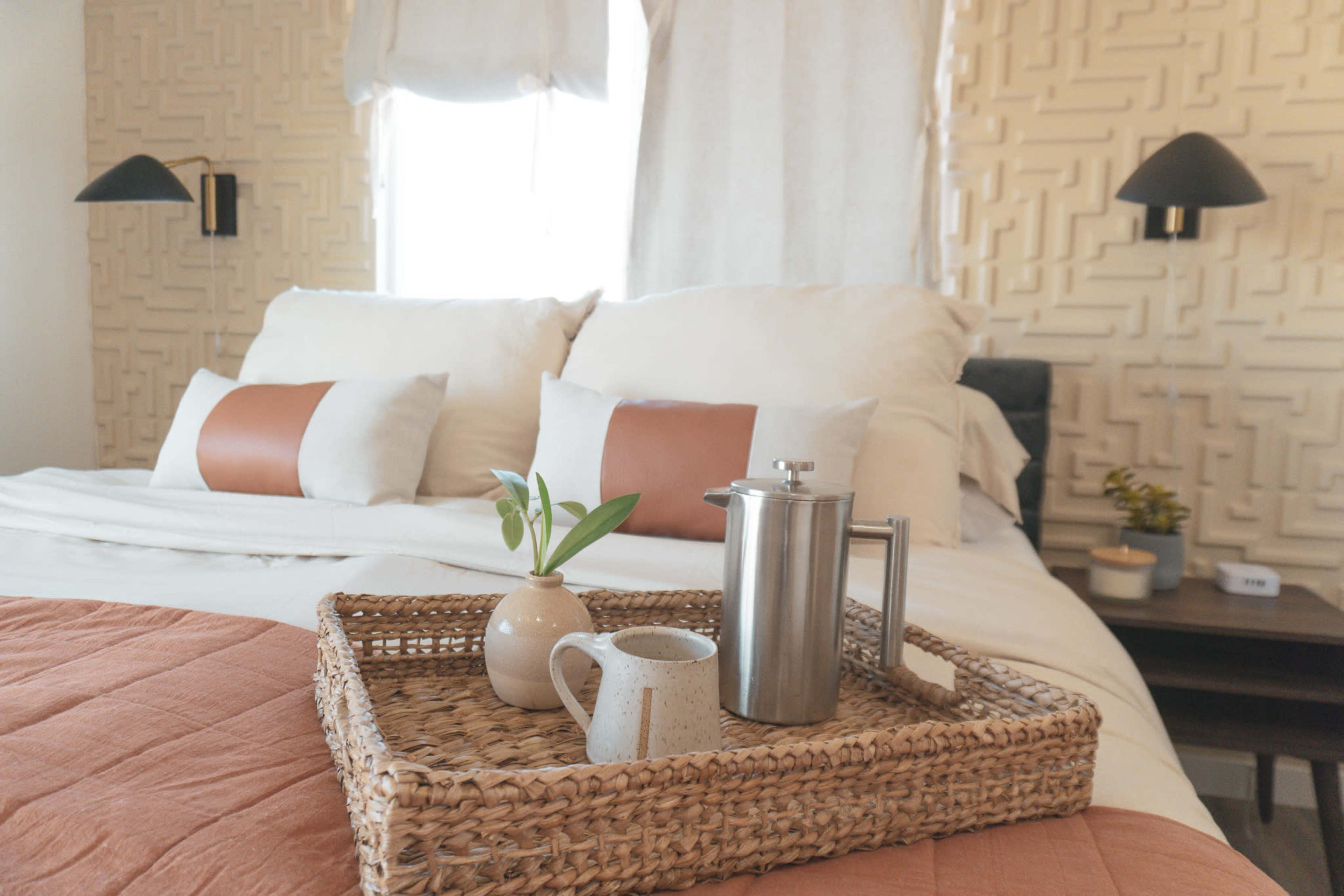 The image shows a neatly arranged bed with white linens, decorative pillows, and a tray holding a coffee pot, a small pitcher, and a plant.