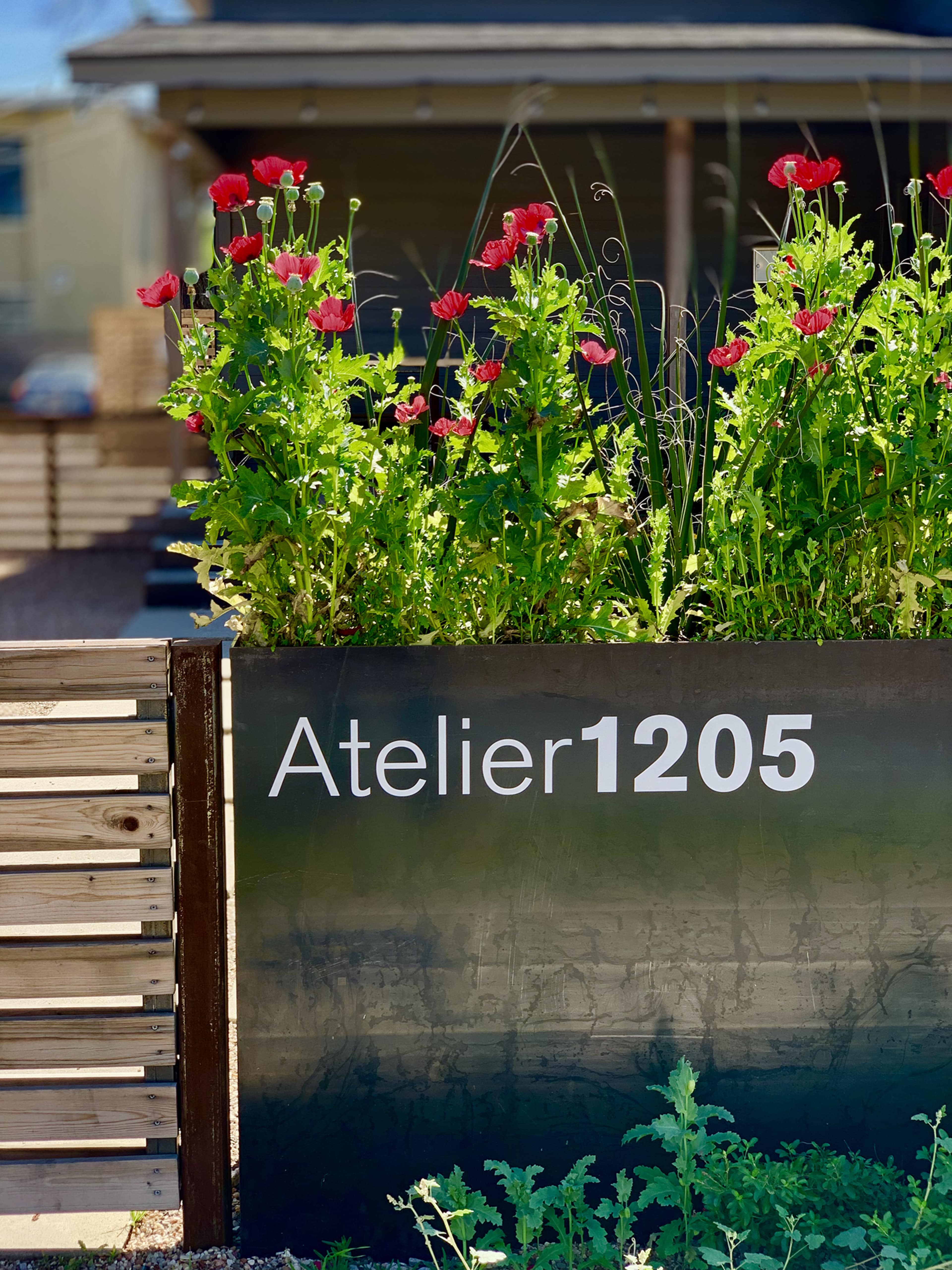 A flower bed with red blooms is situated in front of a sign that reads "Atelier 1205."