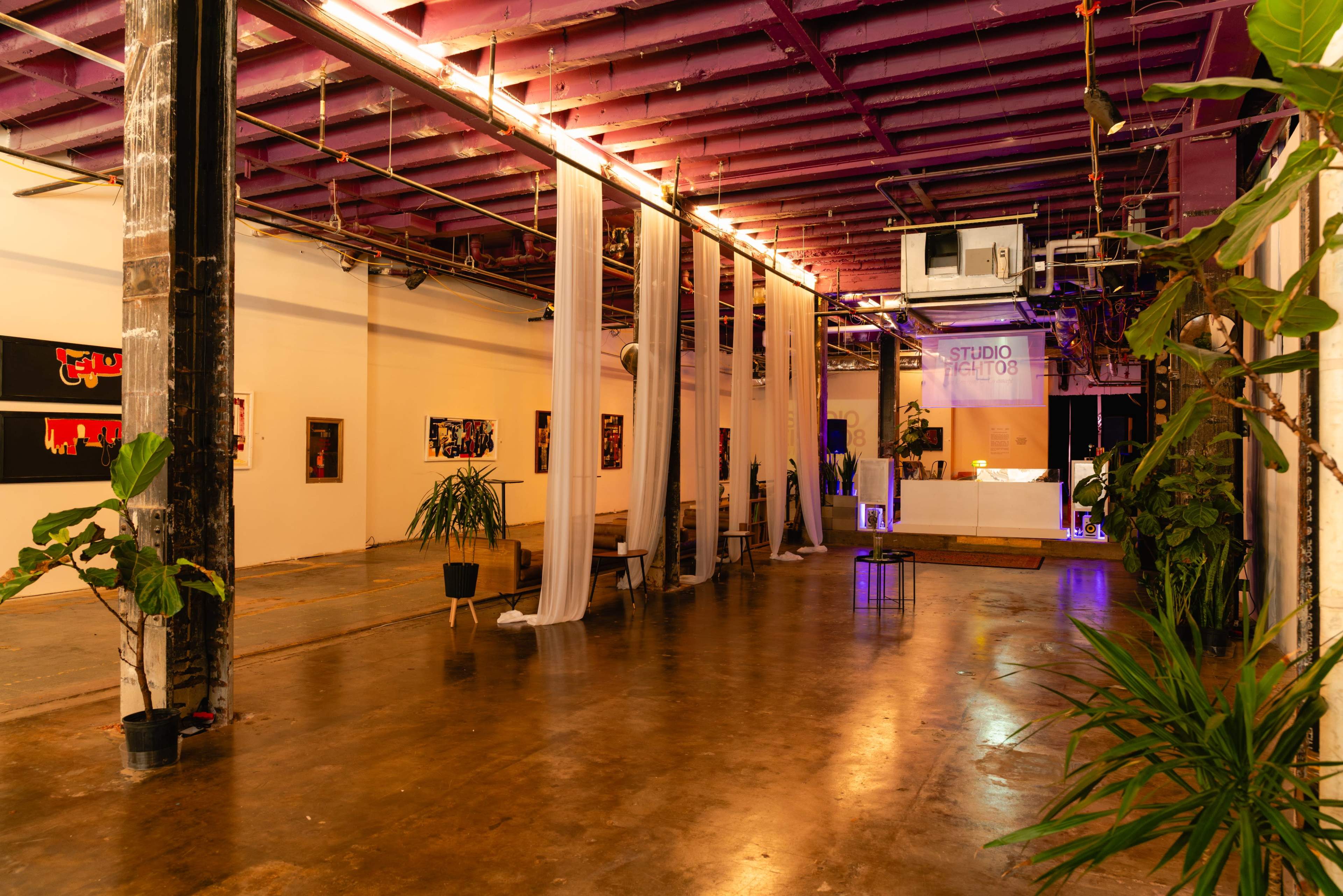 The image shows a spacious gallery with exposed brick walls, purple ceilings, and decorative plants, featuring artwork displayed on the walls and soft lighting.
