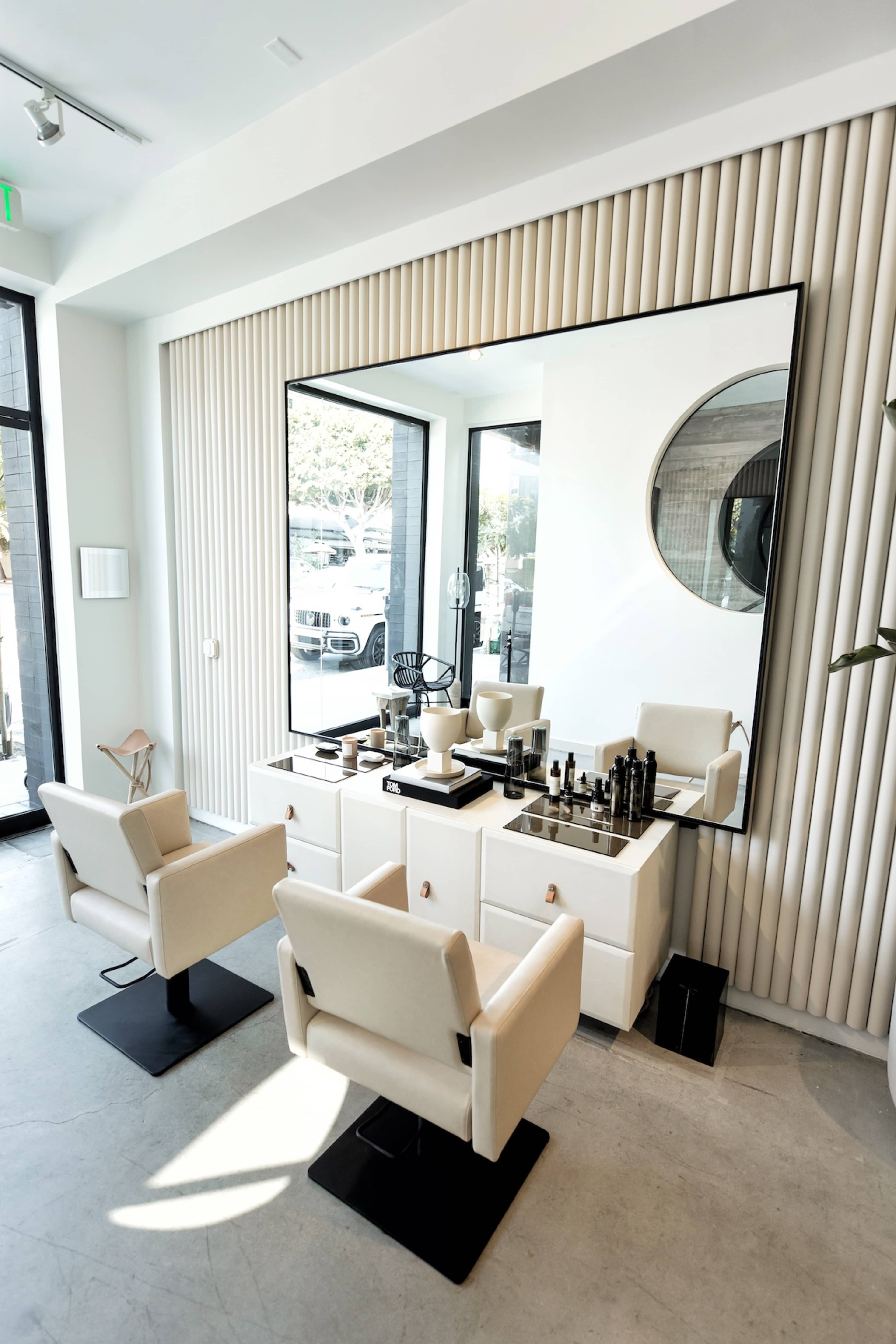 A modern salon interior featuring two white chairs in front of a mirrored vanity with makeup products and a light-colored, textured wall.