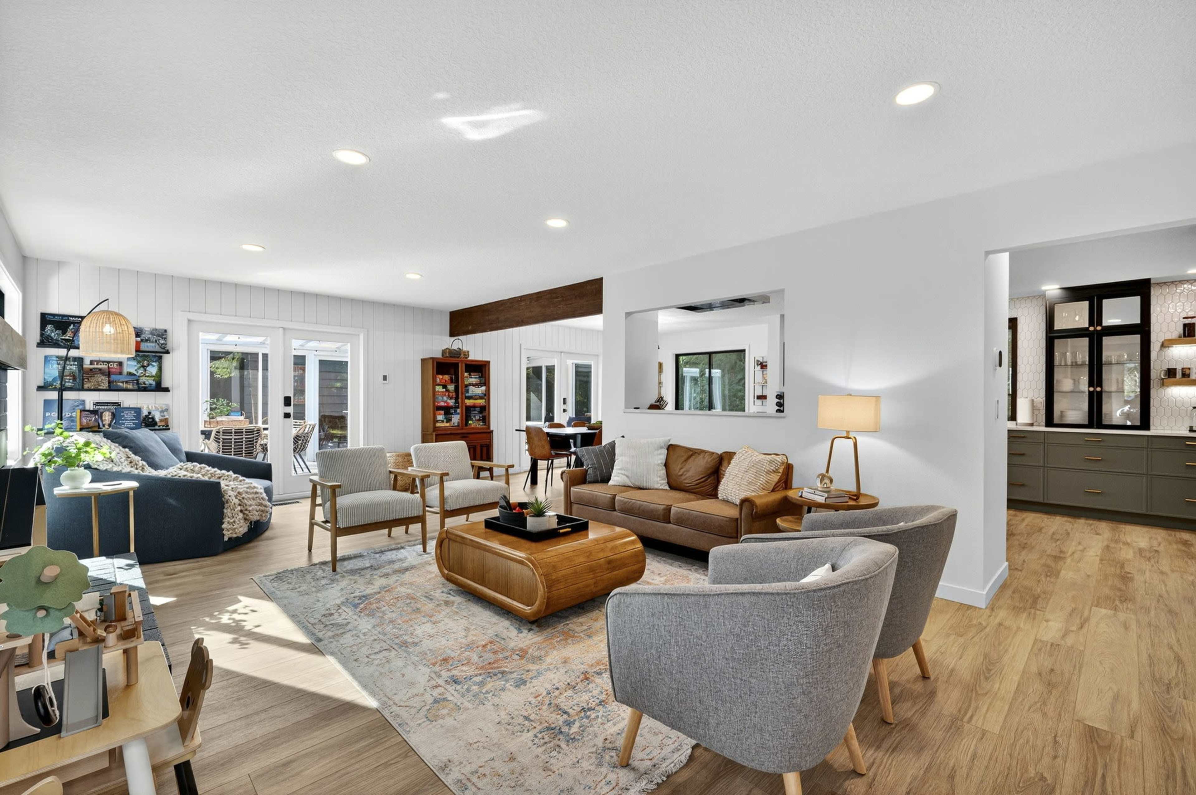 The open living area features a mix of seating, including a sofa and armchairs, with natural light streaming through large windows, and leads into a kitchen area visible in the background.