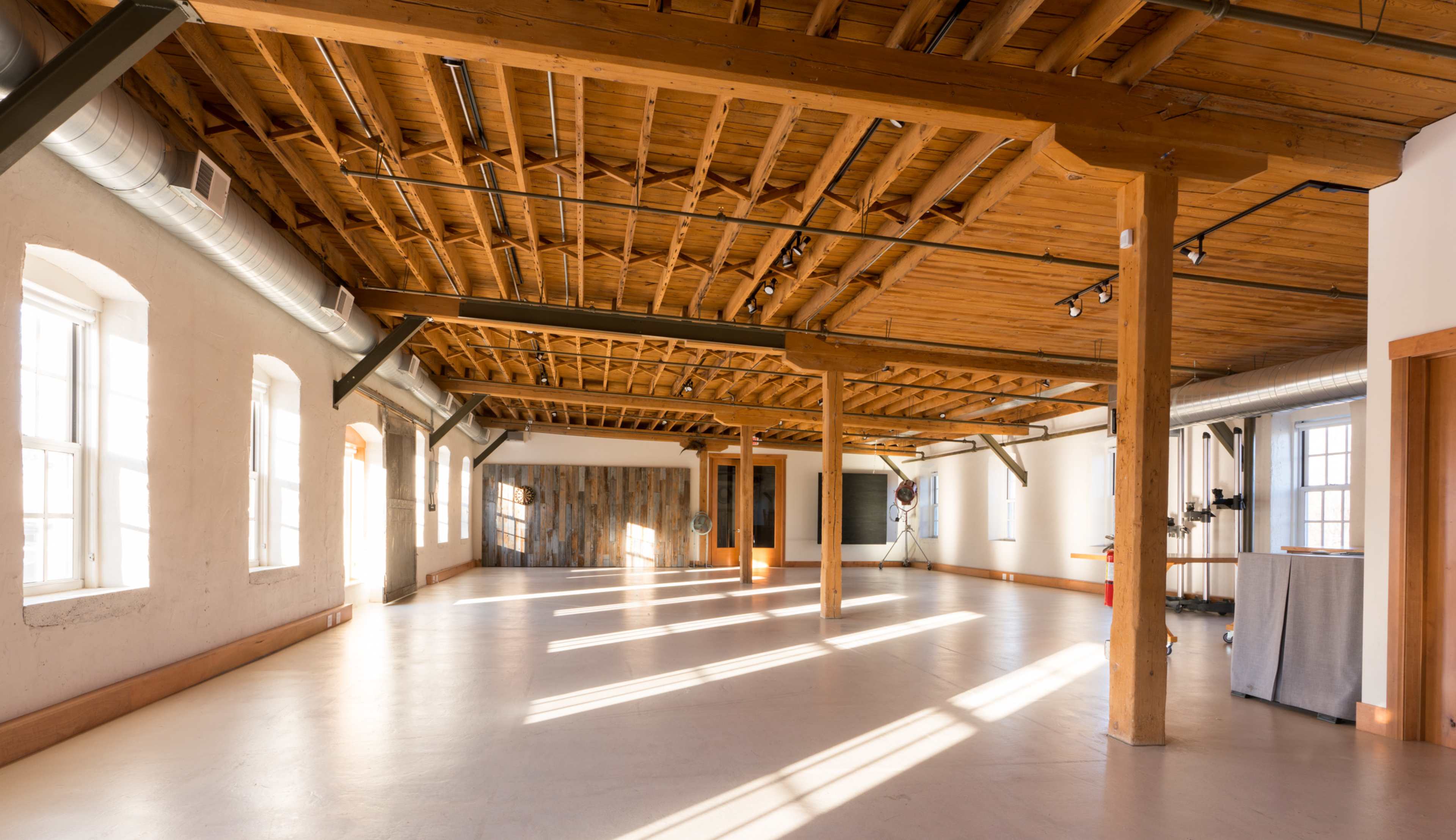 The image shows a spacious, well-lit interior with wooden beams and large windows, featuring concrete flooring and minimalistic decor.