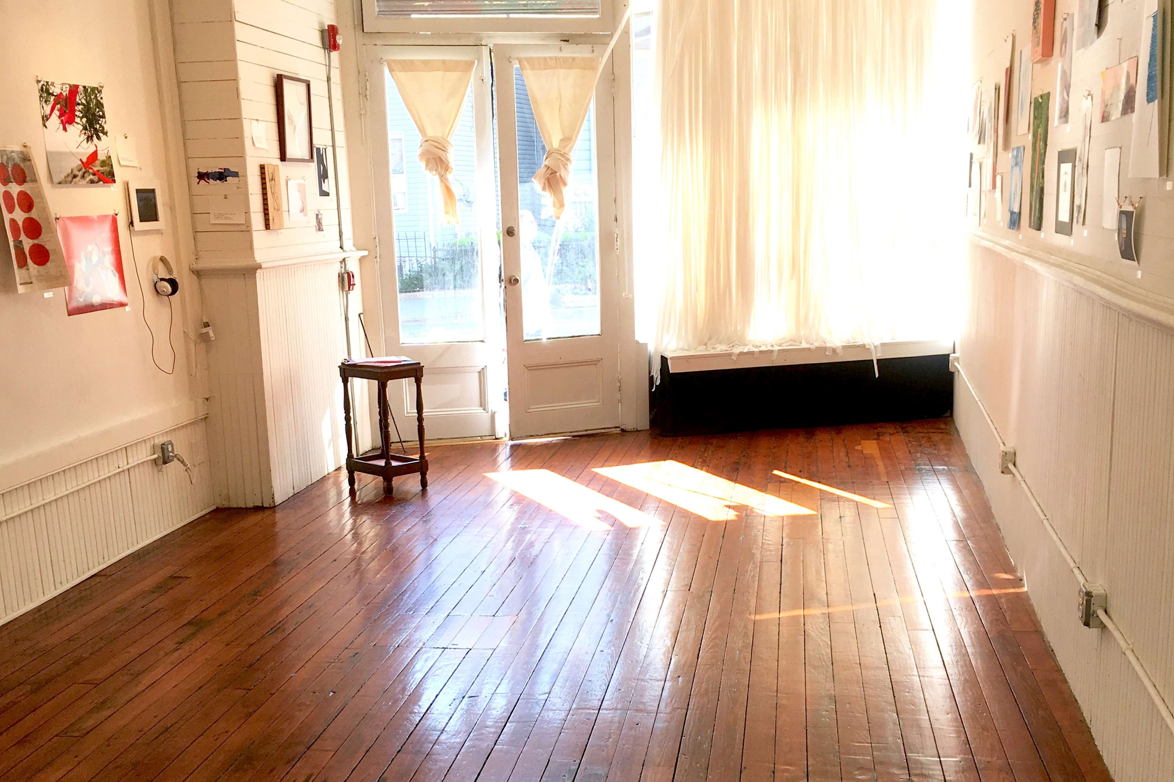 A brightly lit room with wooden floors, a chair against the wall, and large windows draped with light-colored curtains.