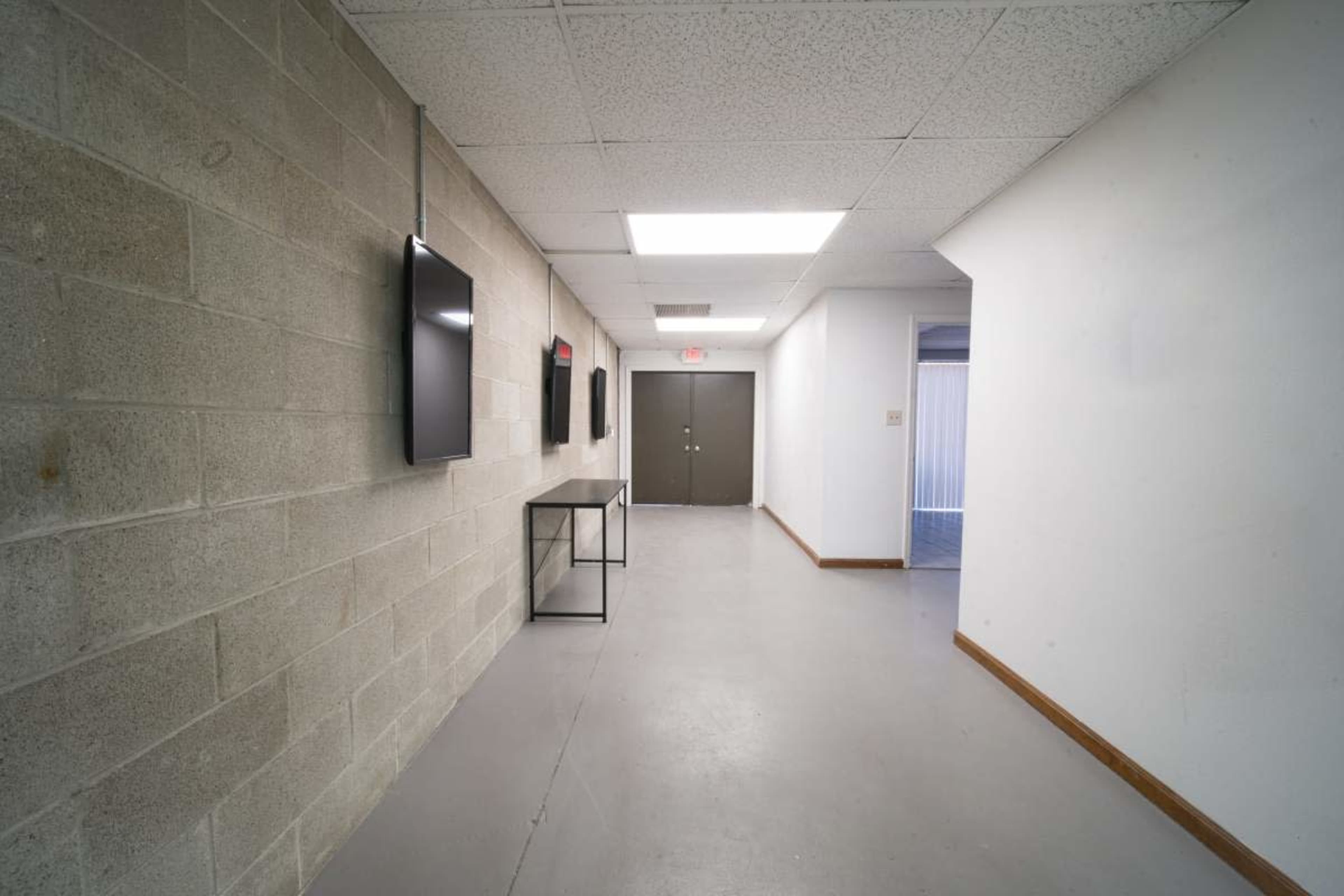 The image shows a long, narrow hallway with concrete walls, two mounted televisions, and a closed door at the end.