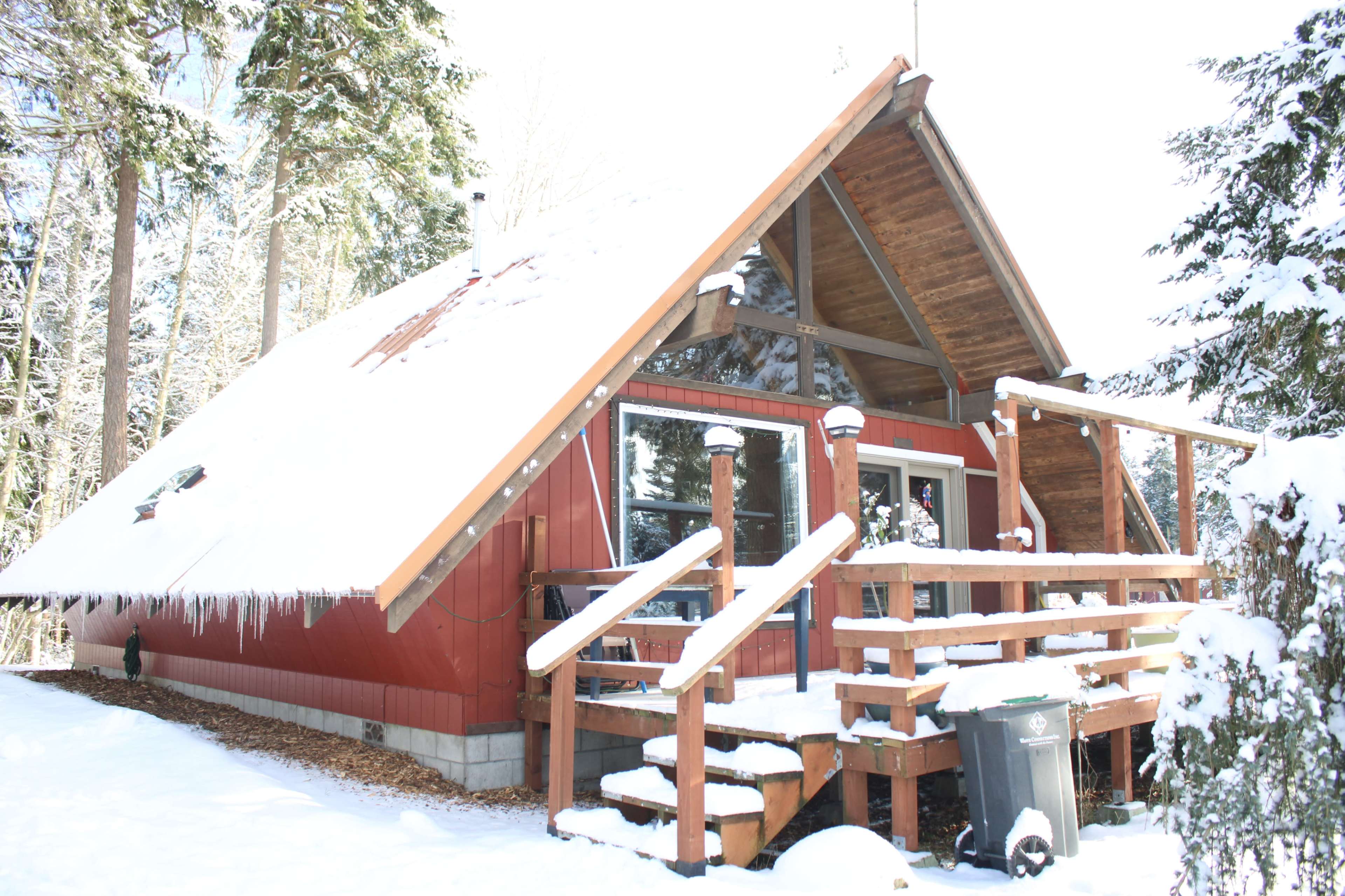 A red A-frame cabin with a snow-covered roof and wooden deck is surrounded by trees in a winter landscape.