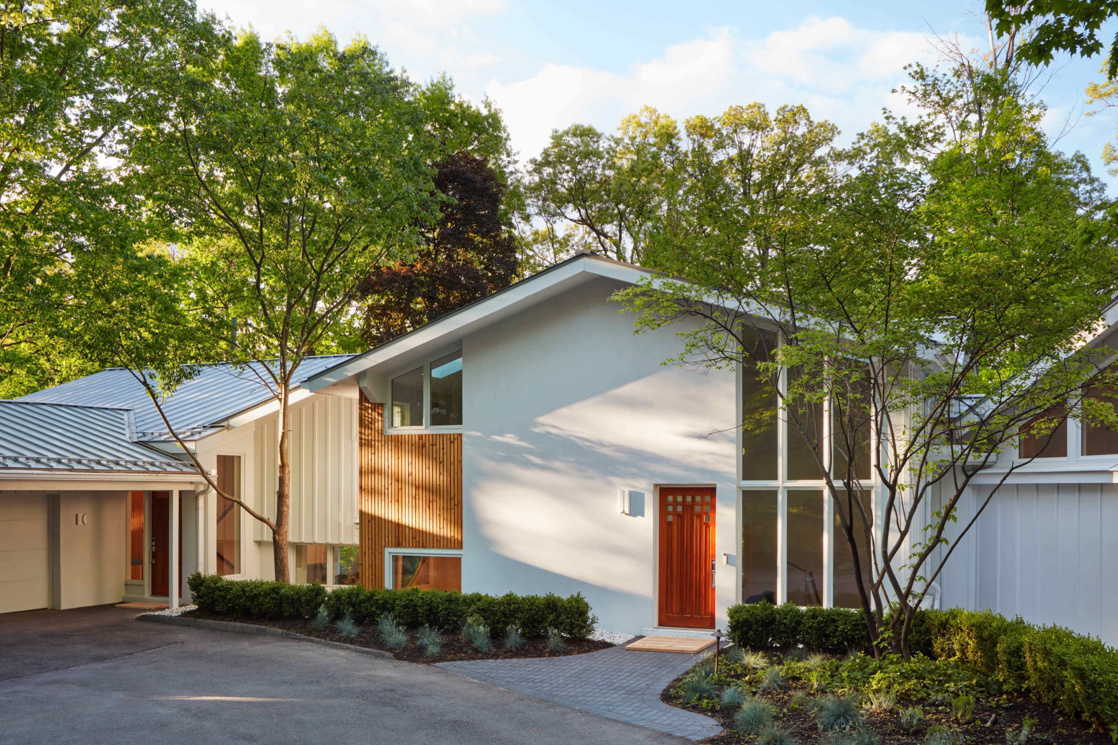 A modern house with a mix of white and wooden exterior is surrounded by greenery and features large windows and a distinct entryway.
