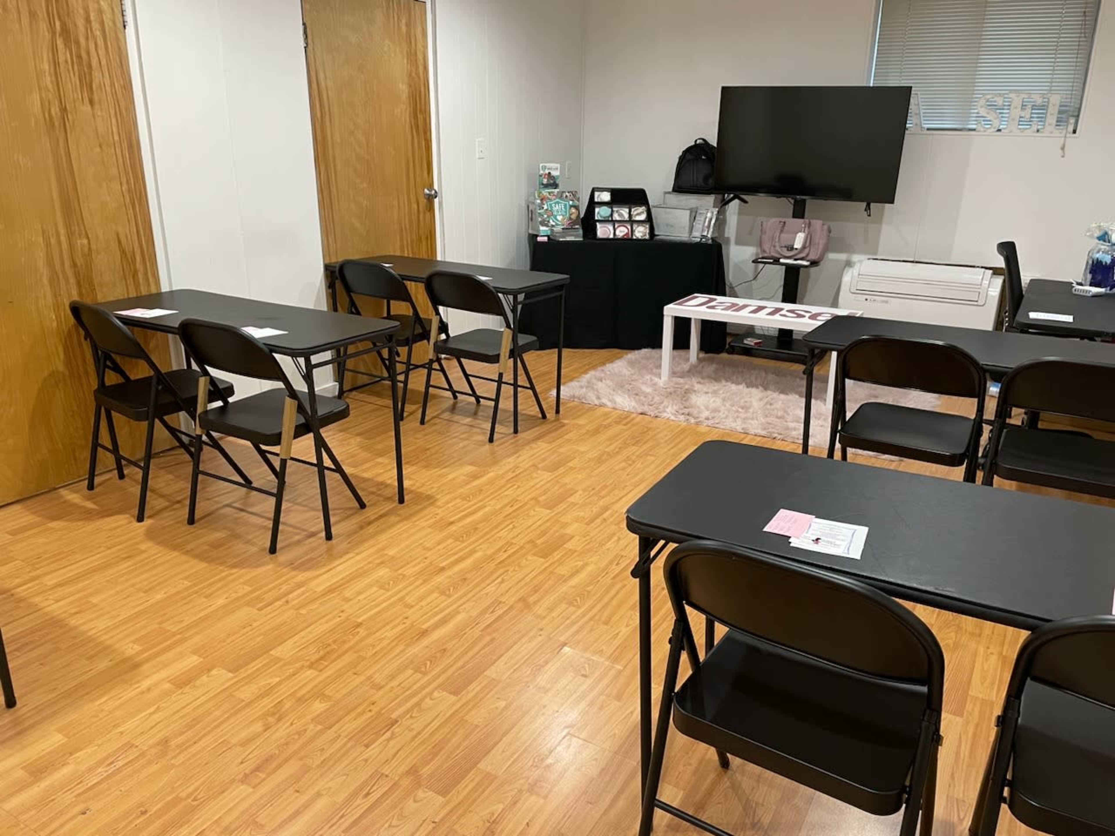 A classroom setup with black folding tables and chairs arranged facing a television in a room with light-colored walls and wooden floors.
