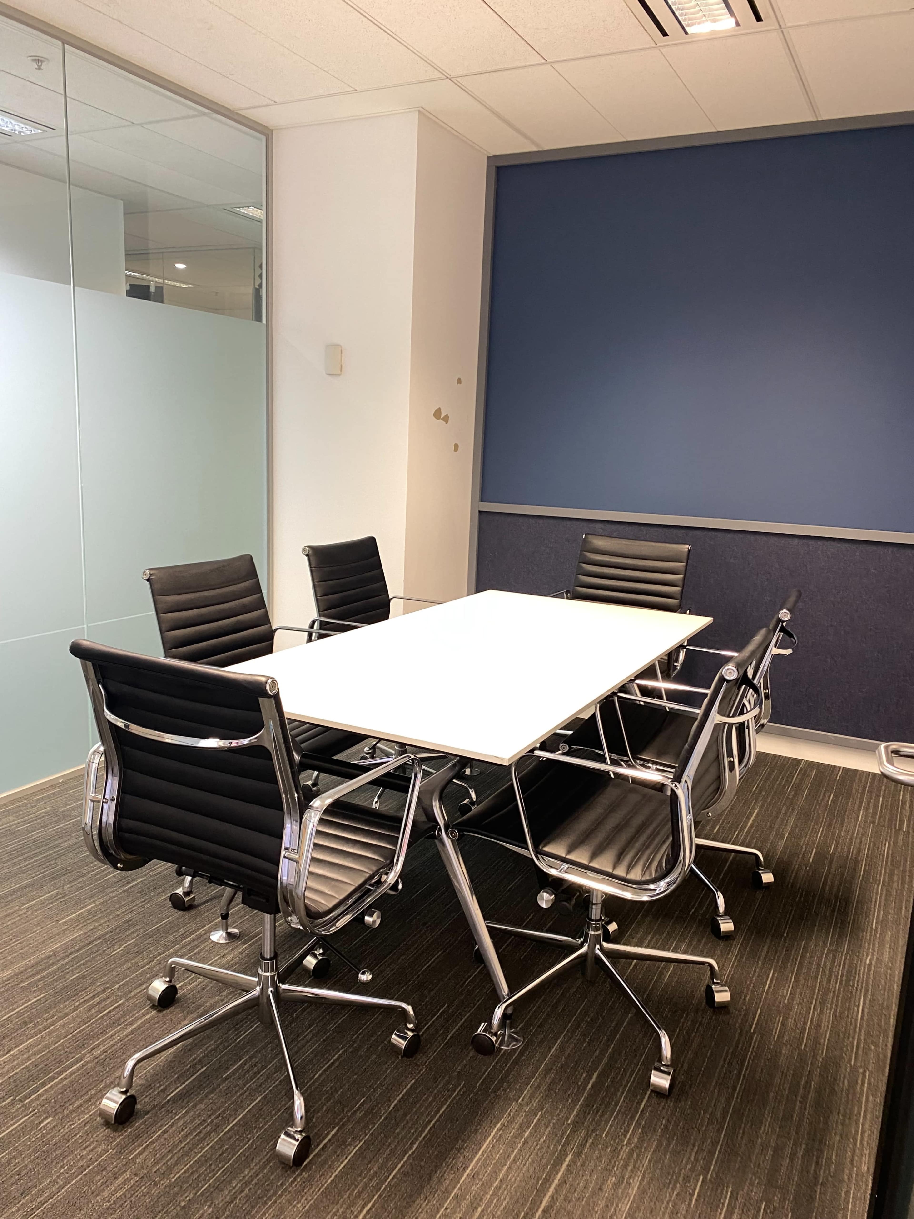 A modern conference room features a white tabletop surrounded by six black rolling chairs on a dark carpet.