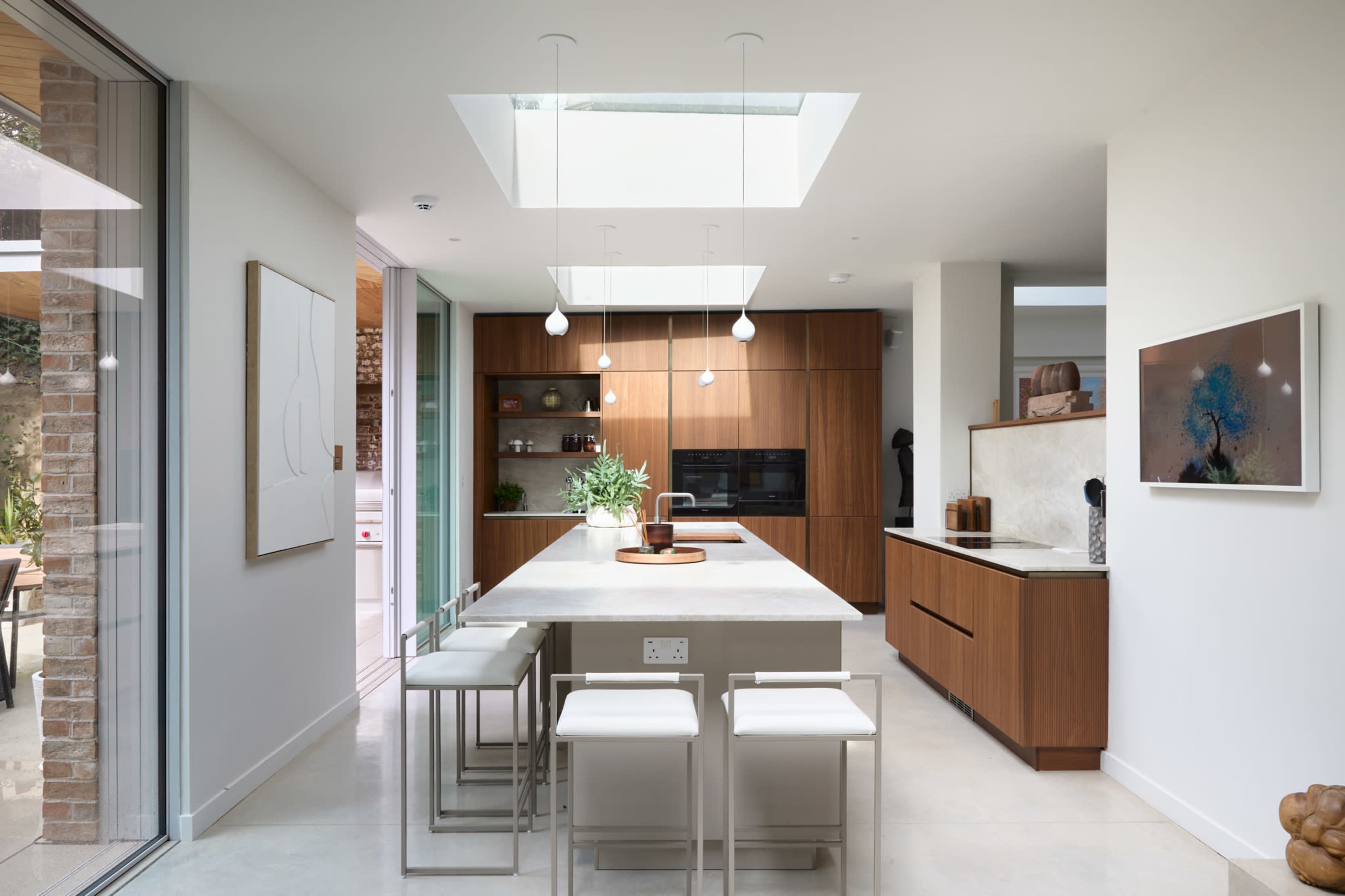 A modern kitchen features a central island with a stainless steel countertop, white bar stools, wooden cabinetry, and a skylight above.